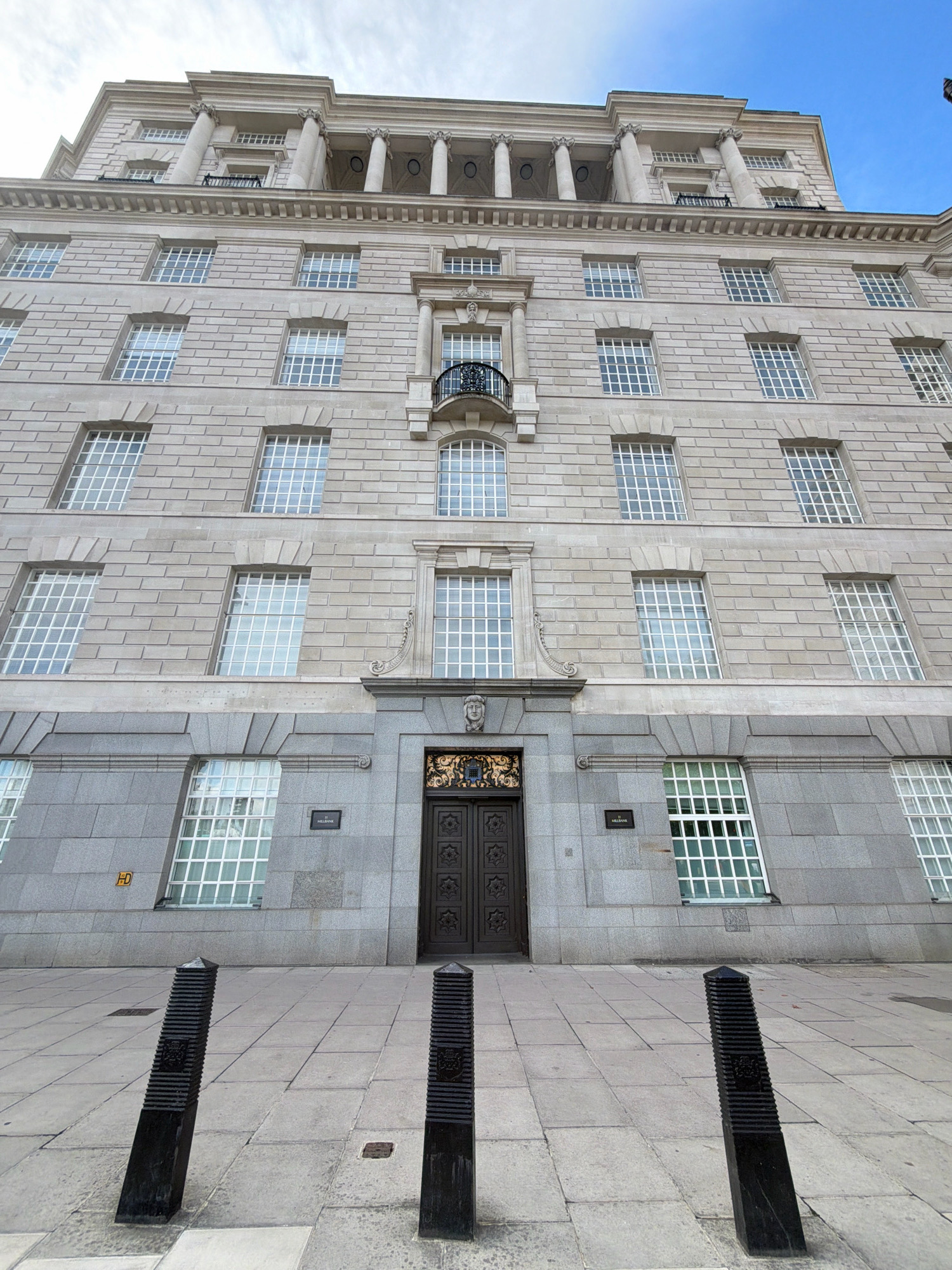 The image shows the exterior of a large, ornate building with a stone facade. The building has multiple floors of windows, many of which are divided into a grid pattern. A dark-colored double door is visible in the center of the ground floor, flanked by decorative stonework. Three black bollards are positioned in the foreground on a paved sidewalk. The sky above is clear and blue. A small sign on the building reads "11 Millbank". This is the exterior of the MI6 Headquarters, also known as SIS Building, located at 85 Vauxhall Cross, London SE1 2UH, United Kingdom.