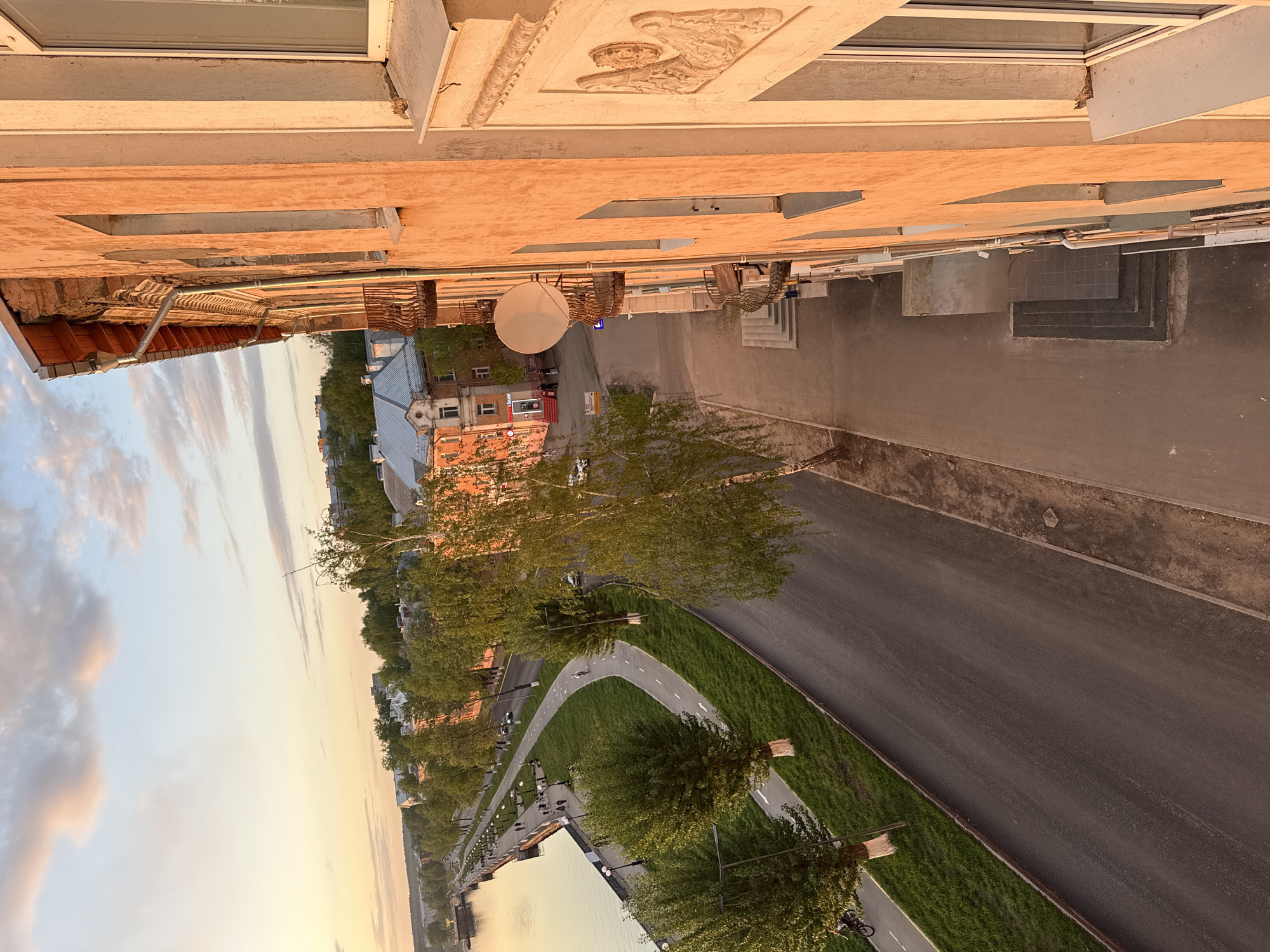 The image shows an elevated view, potentially from a building, overlooking a street in Votkinsk, Russia, possibly in the evening given the lighting.
**Environment:** A wide road runs alongside a grassy embankment and a body of water. A pedestrian/bicycle path parallels the road, closer to the water. Trees line the embankment and path. Buildings are visible on both sides of the road. The sky is light with clouds.
**Objects:** The foreground includes parts of the building from which the picture is taken, like the facade. A satellite dish is mounted on the building, overlooking the street.
**Buildings:** The buildings exhibit diverse architectural styles, with some featuring intricate details. One building is painted in multiple colors.
**People/Activities:** Figures are visible on the bicycle path, appearing to be pedestrians and cyclists. Vehicles are on the road.
In conclusion, the image depicts a street scene in Votkinsk with a road, pedestrian path, waterfront, and a variety of buildings. The scene appears to be a recreational area with people engaged in outdoor activities.