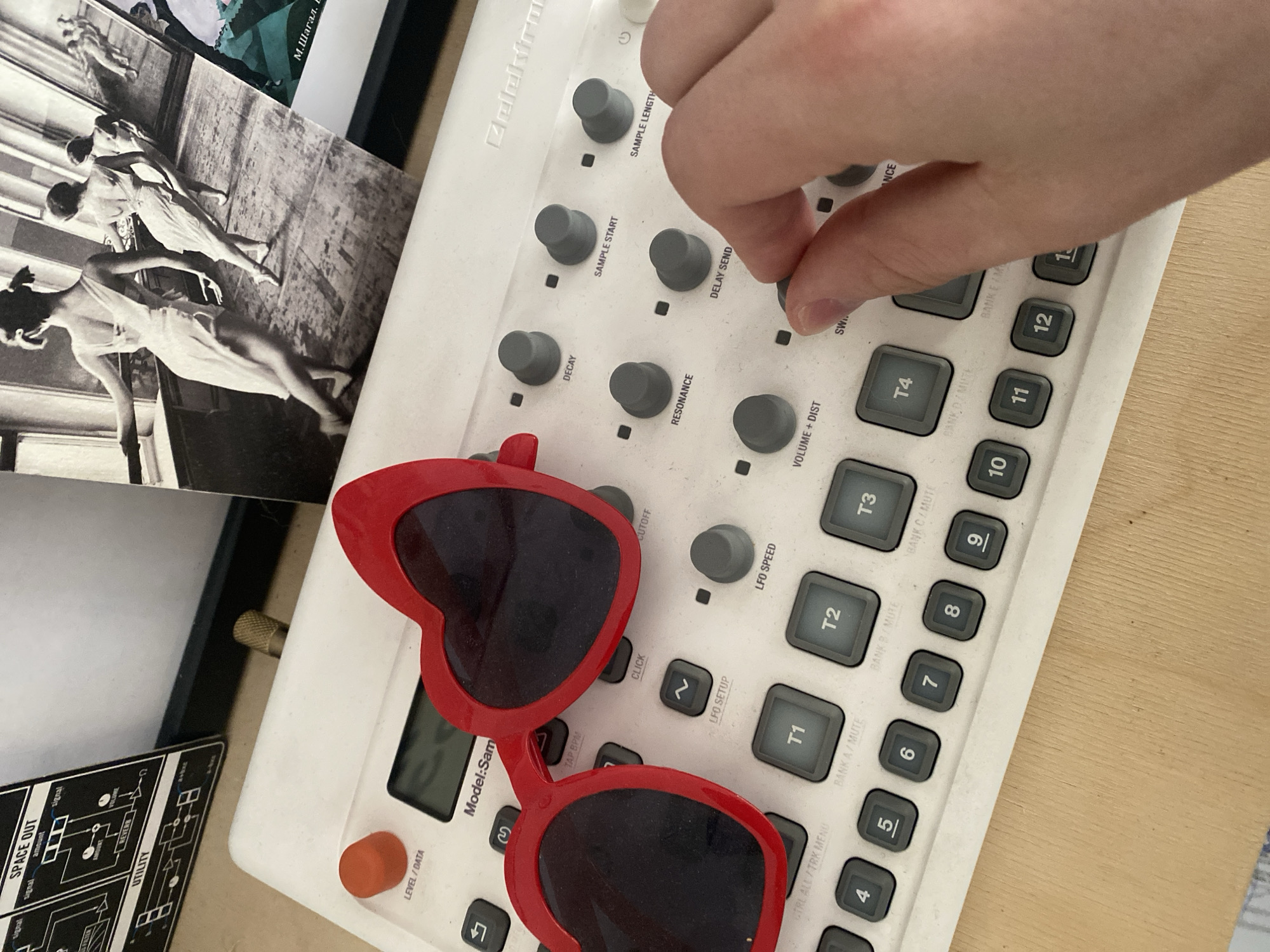 The image shows a person's hand adjusting a knob on a white Elektron Model:Samples synthesizer. A pair of red heart-shaped sunglasses rests on the synthesizer's surface. A black and white photograph depicting dancers in motion and a diagram of electronic signal paths are present in the periphery. The photograph displays the Cyrillic letters “М.Шагал”. The location in the city of Rusanovo, Russia is not determinable from the provided image.