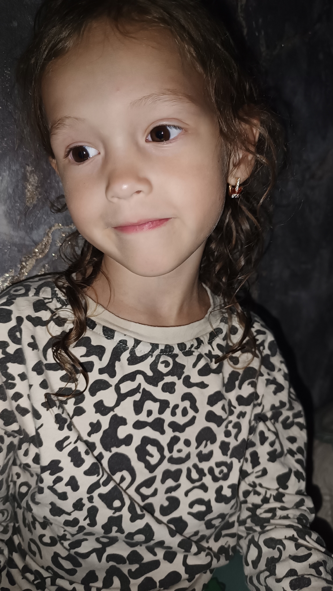 A close-up, eye-level shot shows the upper body of a young girl with curly brown hair and brown eyes, looking slightly to her left. She is wearing a beige shirt with a black leopard print and a small gold and red earring in her right ear. Her mouth is slightly open as if she is about to speak, and her left cheek is gently pressed against her hand. The background is dark and indistinct, with a textured, possibly gray wall to the left. There is no indication of the city of Karsha, Russia, or any specific urban environment.