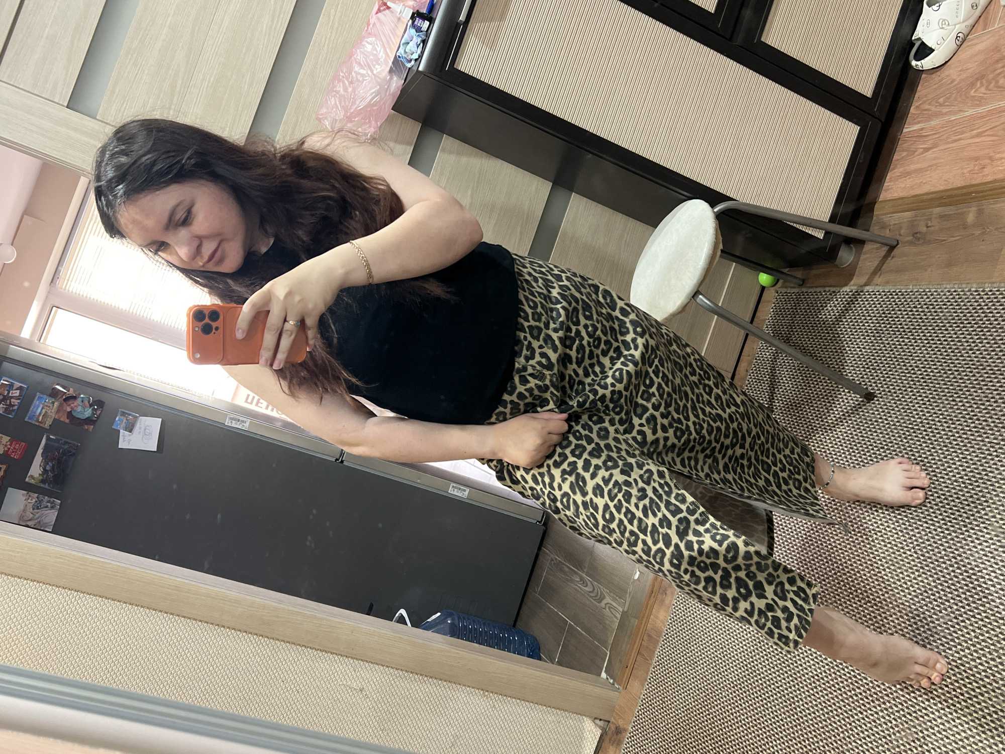 An indoor mirror selfie taken in Noviy, Russia, shows a woman with long, dark, wavy hair, wearing a black sleeveless top and leopard print trousers. She holds an orange smartphone to capture her reflection. A gold-colored braided bracelet adorns her right wrist, and a ring is visible on her left ring finger. Her bare feet are placed on a patterned beige and brown rug.
The setting features light wooden flooring, a dark brown cabinet or wardrobe with light, ridged panels, and a white, round-seated stool with a metal frame. On top of the cabinet, a pink plastic bag and other small items are present. In the mirror's reflection, a dark refrigerator or large appliance is visible on the left, decorated with several magnets, one of which depicts an engagement/wedding ring. A blue plastic container rests on the floor near the refrigerator. A white slipper with the brand "GUCCI" printed on it is seen on the wooden floor in the upper right. Natural light from a window illuminates the scene, indicating daytime. The woman's gaze is directed towards her phone as she takes the picture.
