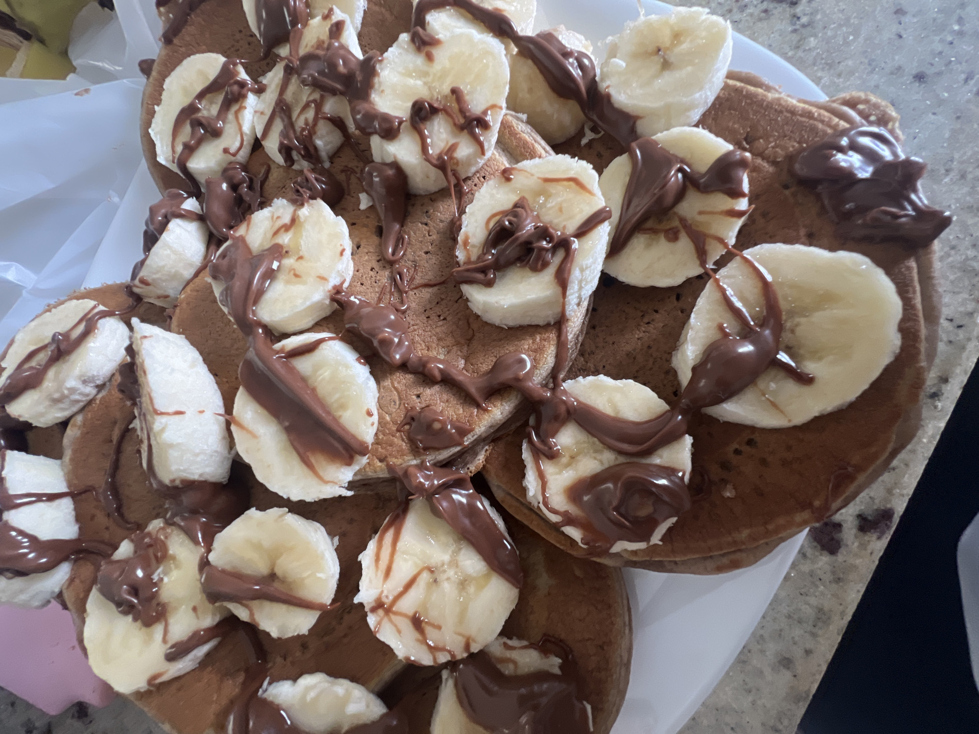 A top-down shot shows a stack of pancakes topped with sliced bananas and chocolate sauce. The pancakes appear to be on a white plate, which is set on a countertop or table with a speckled grey and white surface. To the left, a white plastic bag is partially visible. The scene is well-lit, allowing for clear observation of the food's texture and arrangement.