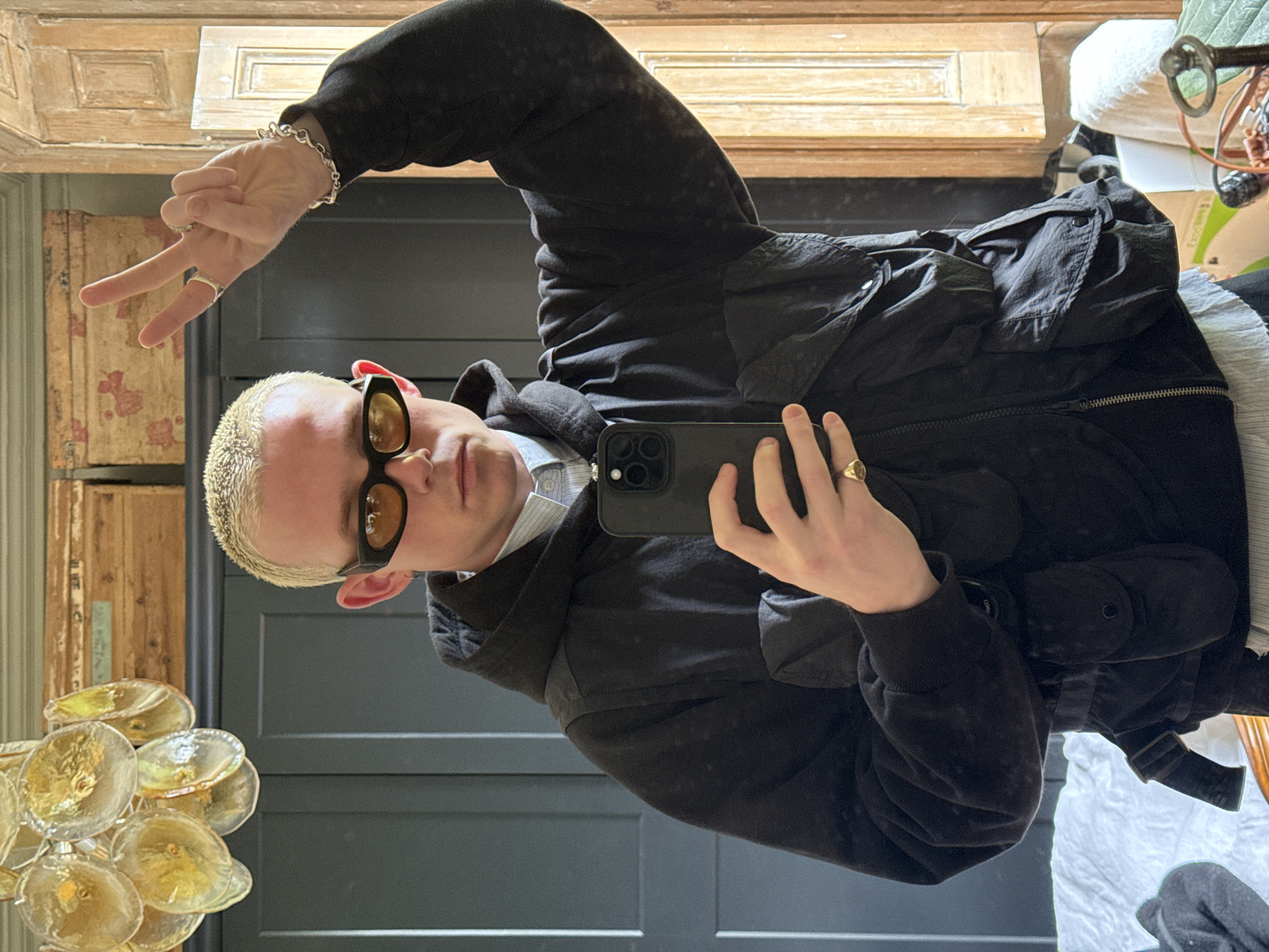A young person with short, bleached blonde hair takes a mirror selfie. They wear dark-framed sunglasses with amber lenses, a dark grey hooded jacket over a light blue striped shirt, and a black utility-style vest with pockets and straps. On their left hand, adorned with a silver chain bracelet and a silver ring, they make a peace sign. Their right hand holds a black smartphone with a triple camera array, displaying a gold-toned signet ring on their pinky finger.
The background features a dark grey paneled wall or door. To the left, an antique wooden structure, possibly a cabinet or door frame, shows distressed wood and remnants of a red and white floral wallpaper-like pattern. Below the person's left hand, faint text "BLE RUE" and some numbers are visible on this wooden surface. In the bottom-left foreground, part of a decorative light fixture with multiple amber-colored, disk-shaped glass or acrylic elements is visible. The upper background shows a lighter wooden ceiling or wall reflecting in the mirror. To the far right, indistinct items, including a white box with "packaging" partially visible, are present. The image captures a casual indoor moment, likely in a home setting in London, United Kingdom.