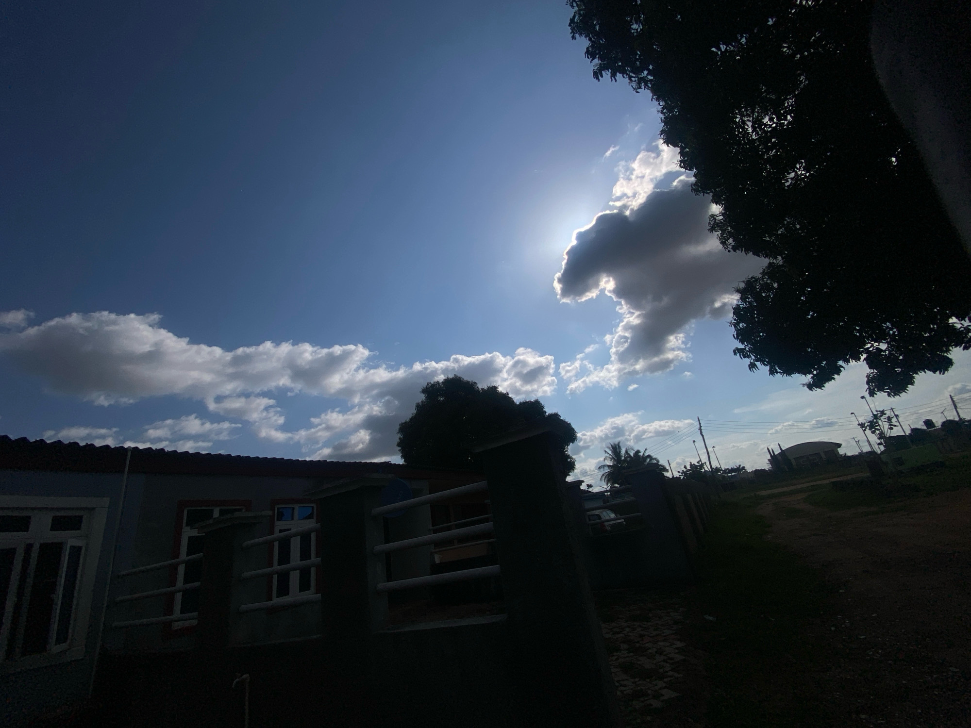 The image shows a cloudy sky with the sun peeking through the clouds, casting a bright glare. In the foreground, there is a dark silhouette of a tree on the right, and a fence or wall with a house partially visible on the left. The house has windows and a red-tiled roof. The ground appears to be a dirt path. The scene suggests a residential area in Barden Yana, Nigeria, likely outdoors during the daytime. There are no people or animals visible in the frame. The overall lighting is mixed, with bright sunlight in the sky and dark shadows in the foreground.