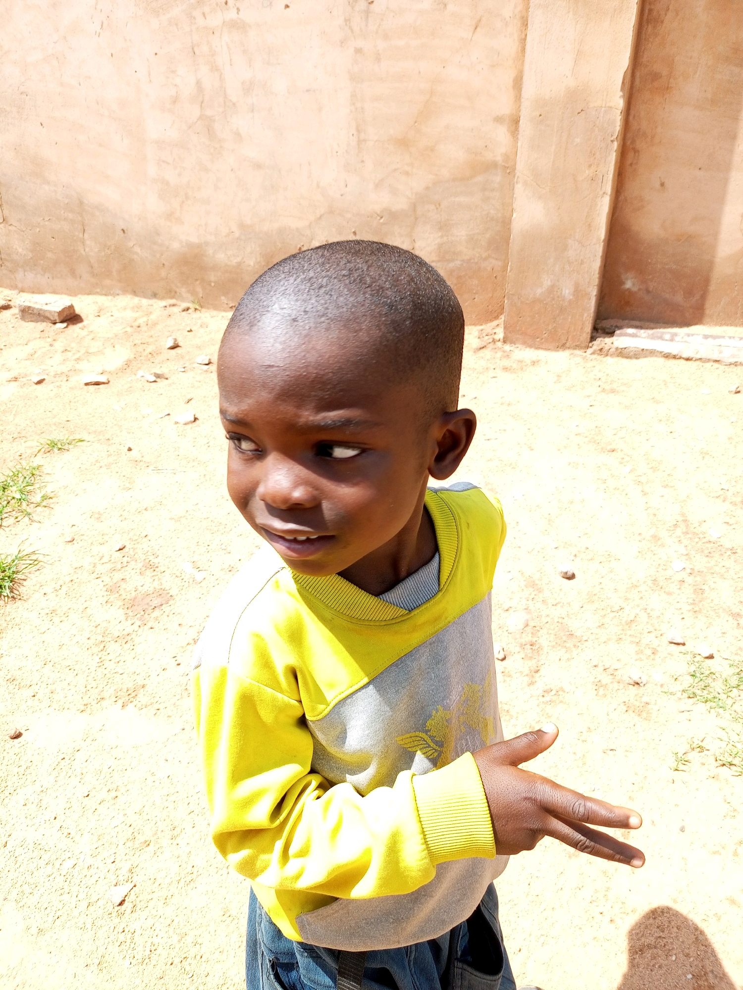 A young boy stands on a dirt ground, looking to his left. He has dark skin and a short haircut. He is wearing a yellow and gray long-sleeved shirt and blue jeans. His right hand is raised, with his index and middle fingers extended, forming a V-shape. The background consists of a tan wall and sandy ground with some sparse vegetation. The scene appears to be outdoors, possibly in a rural or semi-urban area, given the unpaved ground and simple wall construction. The lighting suggests it is daytime, with the sun casting shadows. There is no indication of the city of Tempure, Nigeria in the image.