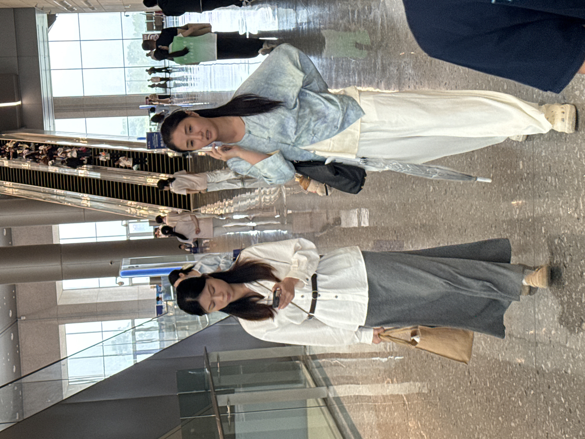 In a brightly lit, modern interior space with polished floors and large windows, two young women walk towards the camera. The woman on the left wears a white long-sleeved shirt with a dark belt and a long grey skirt, carrying a beige tote bag. She is looking down at her phone. The woman on the right wears a pale blue patterned top over a white undershirt and loose white pants. She is talking on her phone and holding a clear umbrella. In the background, an escalator is filled with people, and other individuals are visible in the distance. The overall environment suggests a public building, possibly a transportation hub or a commercial center. The scene is located in the Chongqing High-tech Zone, China.