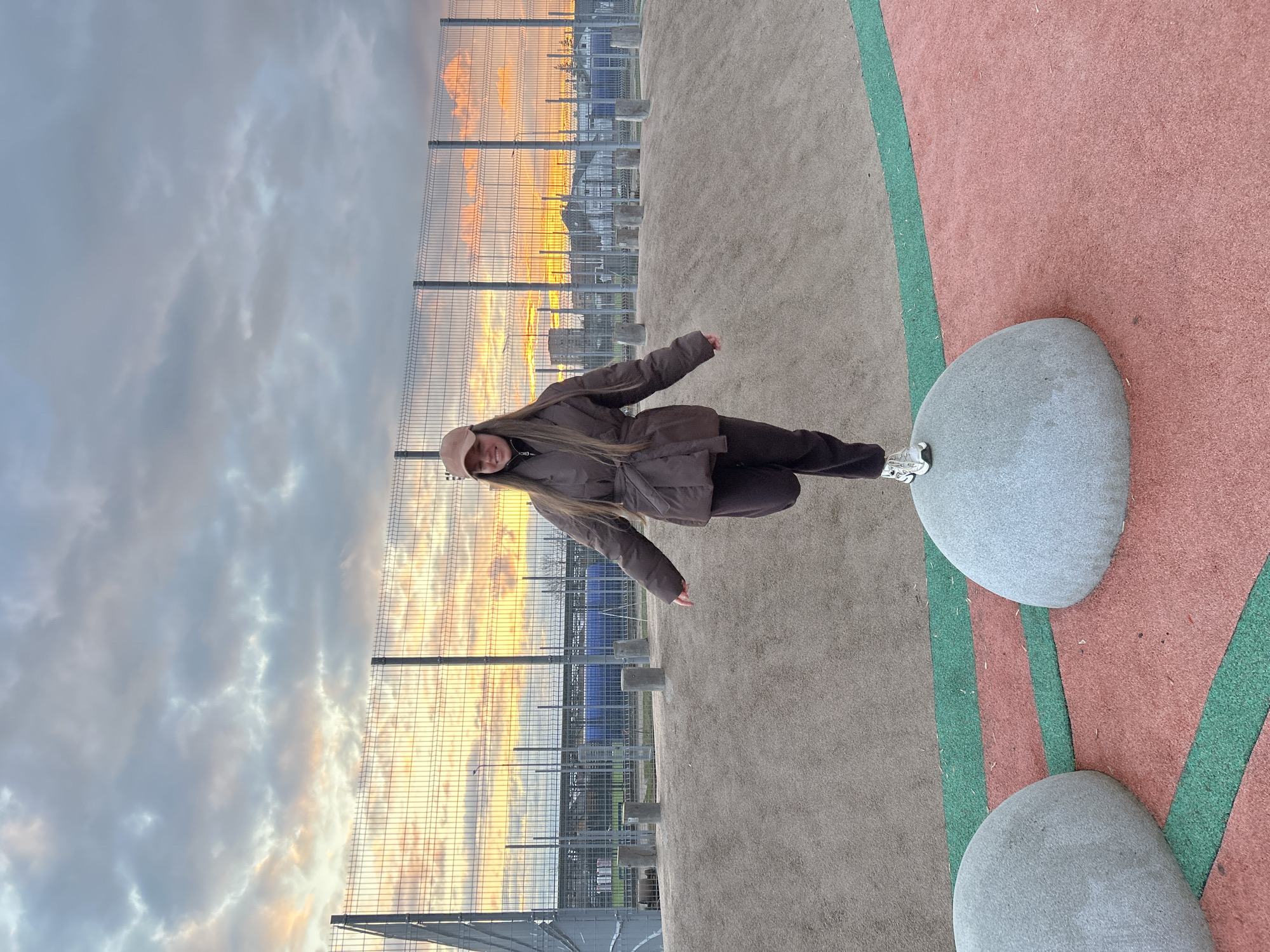 The image shows a young woman standing on a gray stone in a play area. She is wearing a brown jacket, brown pants, a beige hat, and white sneakers. Behind her is a metal fence, beyond which buildings and a colorful sky can be seen. The ground is covered in a rubberized material, with sections colored red, green, and gray. The location appears to be a play area in the city of Klin, Russia.