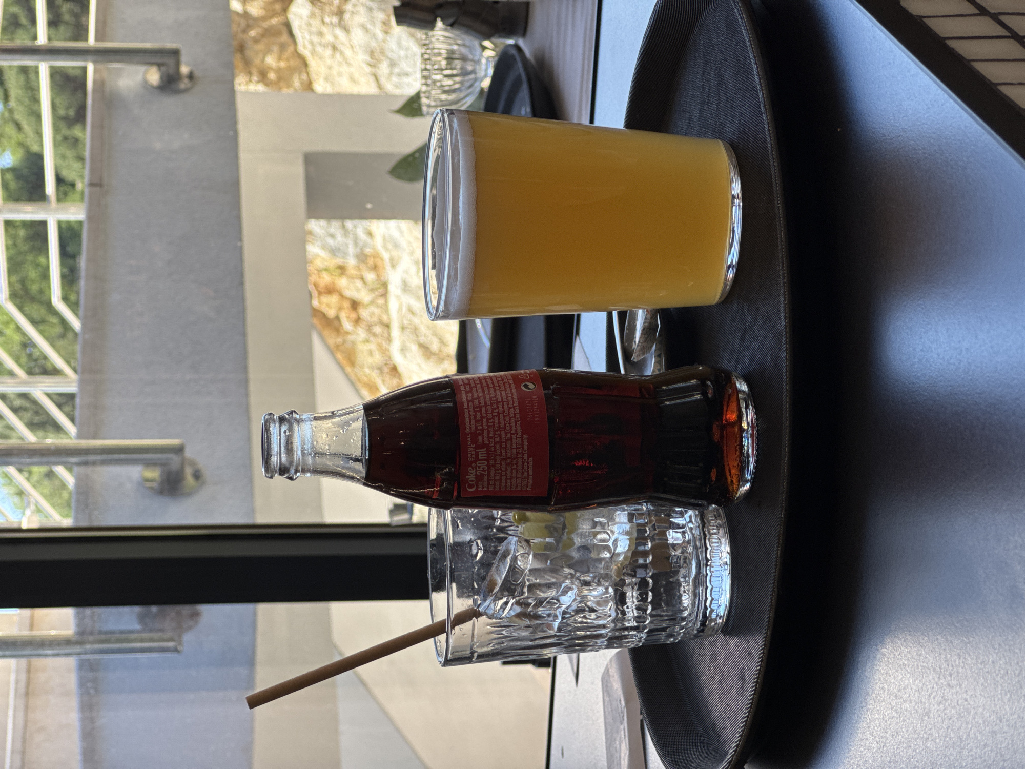 The image captures a black tray on a table with a beverage arrangement. A glass of orange juice stands upright, showing a foamy head. A bottle of Coca-Cola lies on its side, partially obscuring a glass filled with ice and a brown straw. A window in the background reveals trees and a stone wall. The OCR text visible on the Coca-Cola bottle suggests it's a 250 ml bottle containing natural flavors. The text also mentions "Boreti," indicating the scene is located in the city of Boreti, Montenegro.
