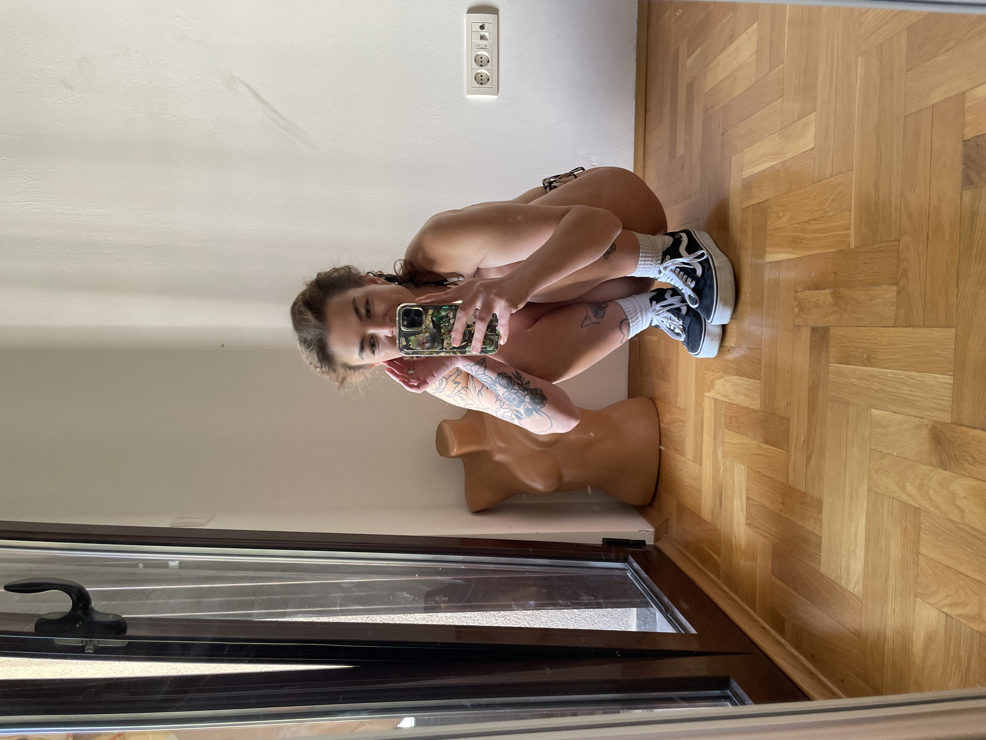 A person with light skin and curly brown hair is crouched down on a wooden floor in front of a mirror, taking a selfie with a mobile phone. The person is wearing sneakers with white socks and appears to be wearing minimal clothing, only visible parts are animal print bikini bottoms. The phone has a camouflage pattern case. The person has tattoos on their arm. A partially visible mannequin torso is next to them. The room has white walls and a European-style electrical outlet. A partially visible window is in the foreground. Based on the style of the room, the outlet, and the parquet flooring, the location could potentially be a domestic interior setting in a city like Boreti, Montenegro, if the image was taken in that location.