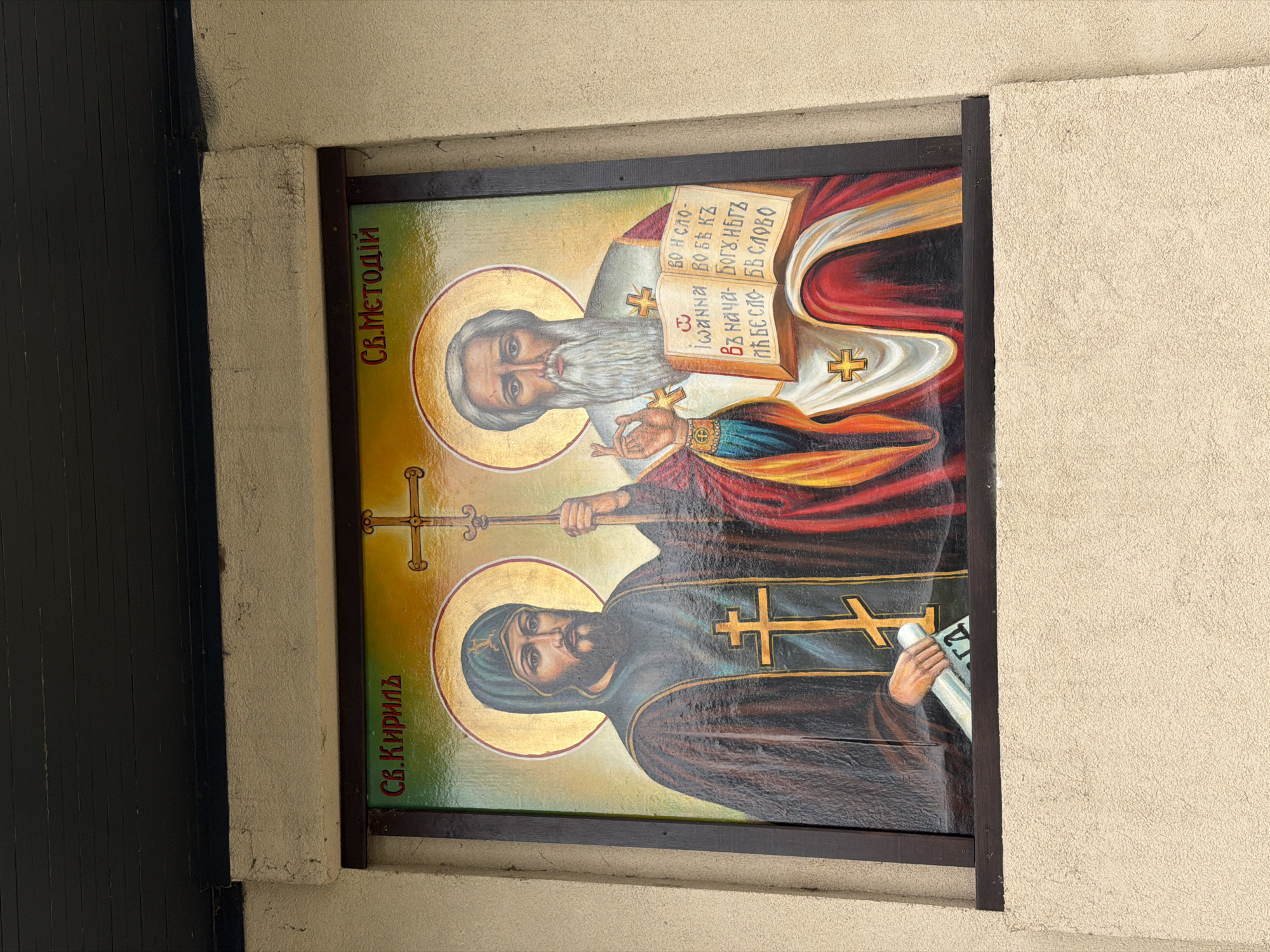 The image depicts a rectangular Orthodox Christian icon or painting of Saints Cyril and Methodius, encased in a dark brown frame and mounted on a light-colored textured wall. The writing in Bulgarian on the left of the icon reads "Св.Кириль" and "Св.Методій," indicating the names of the saints: Cyril at the bottom and Methodius at the top. A partial translation on an open book, held by the saint at the top, reads "во и сло- THE BOБЪ КЪ" and "Въ нача- Богу.НБГЪ." Cyril is depicted wearing dark robes with a gold cross, and holding a scroll. Methodius wears white and red robes, holding a cross. The background is greenish-yellow with golden halos around each of the saint's heads. The image is taken outdoors, as suggested by the natural lighting and the presence of the textured wall, next to a dark-colored door. The city of Blagoevgrad, Bulgaria is not mentioned, so I cannot determine it.