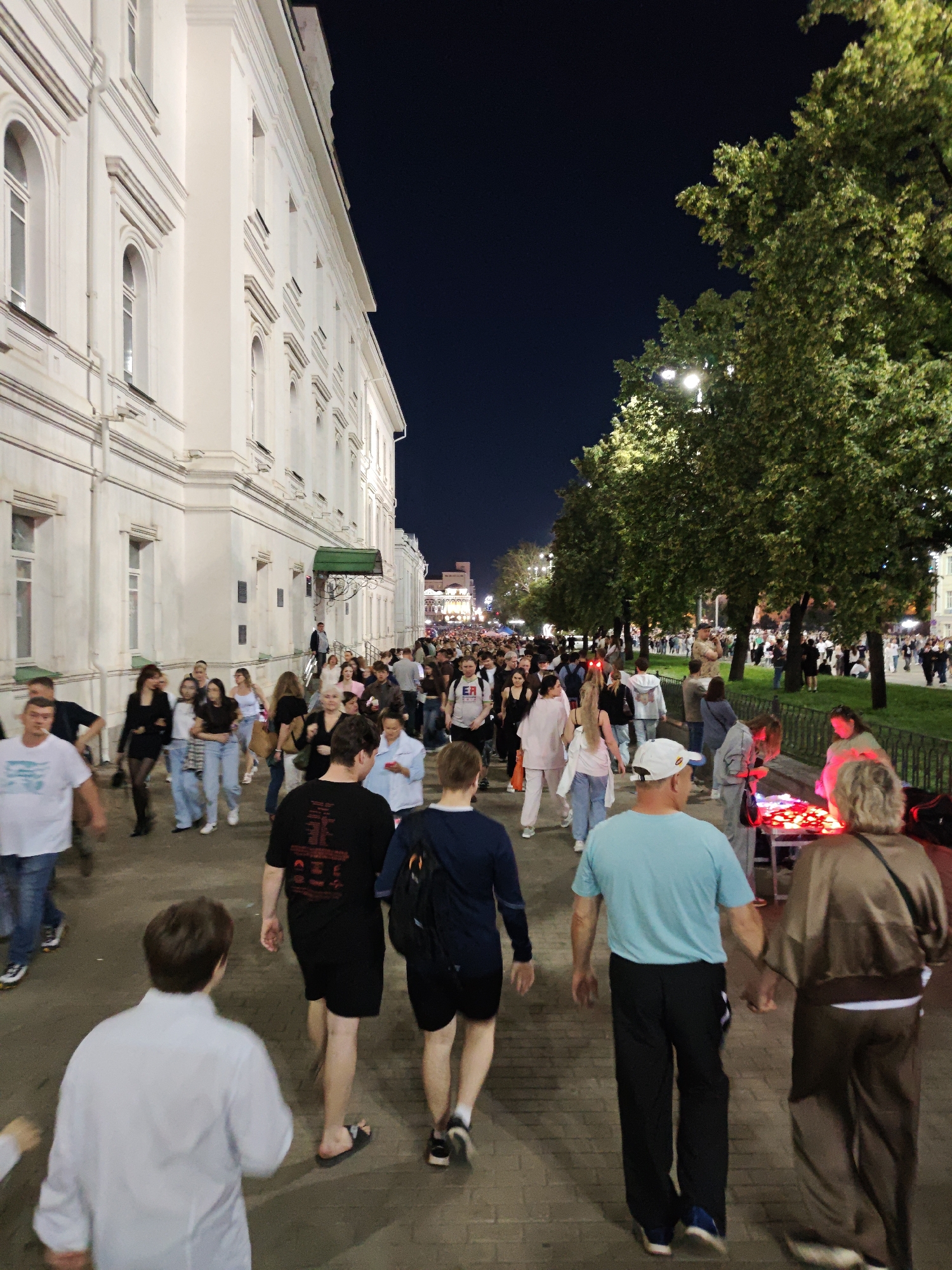 The image depicts a busy pedestrian street scene at night in Yekaterinburg, Russia. A large, white building with arched windows lines the left side of the street, reflecting the ambient light. On the right, mature trees with dense foliage are illuminated by streetlights. A crowd of people are walking along a paved pathway, suggesting a public event or a lively evening promenade.
In the foreground, several individuals are walking away from the camera. A man in a white shirt and blue jeans is on the far left, smoking. To his right, a man in a black t-shirt and shorts walks with sandals. Further right, a man in a blue t-shirt and black pants walks with blue sneakers. He is holding hands with a woman in a brown tracksuit.
In the middle ground and background, the crowd continues, with individuals dressed in casual attire. Some are looking at their phones, while others are engaged in conversation. A stall or table with illuminated red objects is visible on the right side of the path, possibly selling goods. The overall atmosphere suggests a vibrant, public gathering.