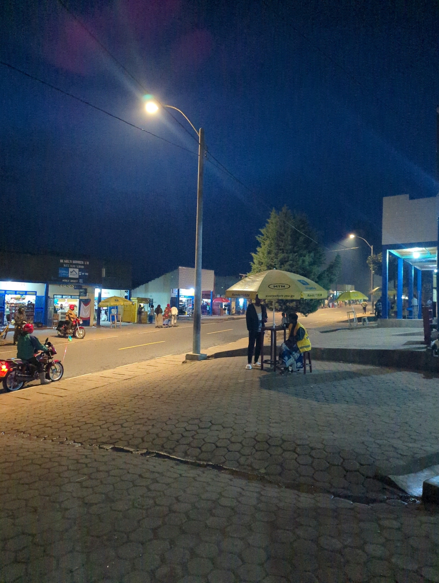 This is a night-time, eye-level, full shot of a street scene in Musanze District, Rwanda. The sky is dark blue, with a street lamp casting a bright light over the road and sidewalk. On the left side of the street, several buildings are visible with illuminated storefronts. A red motorcycle with a rider in a red helmet is parked on the side of the road. Further down the street, another motorcycle is moving, also with a rider.
On the sidewalk, two individuals are standing near a table under a yellow umbrella advertising "MTN". One person, wearing a black jacket and white shoes, stands while the other, wearing a yellow vest and a patterned dress, is seated. There are other people and umbrellas further down the sidewalk, suggesting a marketplace or commercial area. The sidewalk is paved with interlocking hexagonal blocks. The road appears to be asphalt. In the background, trees and more buildings are dimly visible under the street lights.