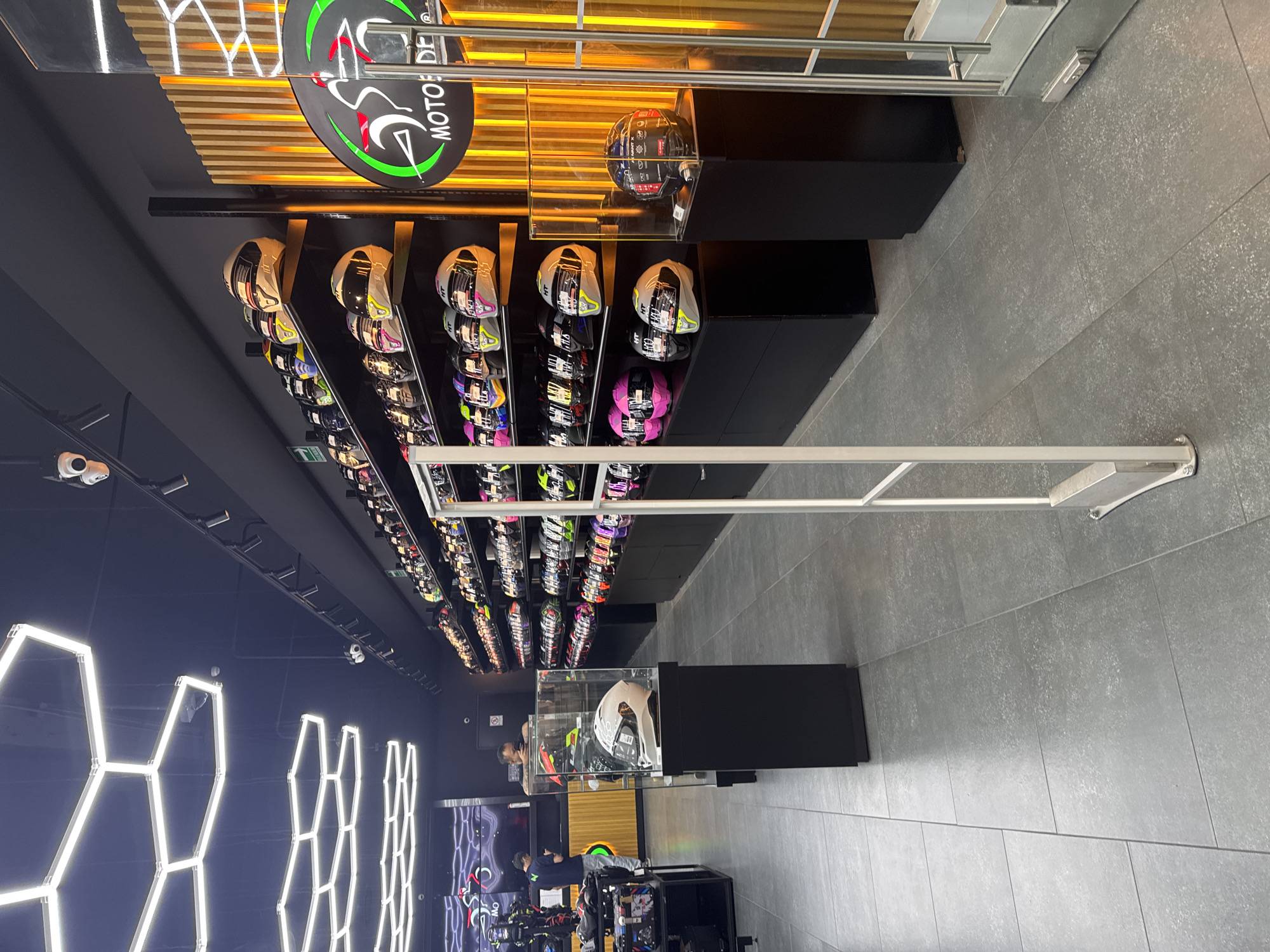 The image shows the interior of a store, likely a motorcycle accessories or helmet shop, in Puebla, Mexico. Numerous motorcycle helmets are displayed on shelves along one wall, extending far into the store. The helmets vary in color and design. A glass display case houses a helmet near the store's entrance. Two people, presumably customers or employees, are visible near the cash register area. The store is brightly lit with a modern design, featuring geometric light fixtures on one wall. The floor is tiled in grey. An anti-theft device is visible in the foreground. A sign with "MOTODE" is seen in the background.