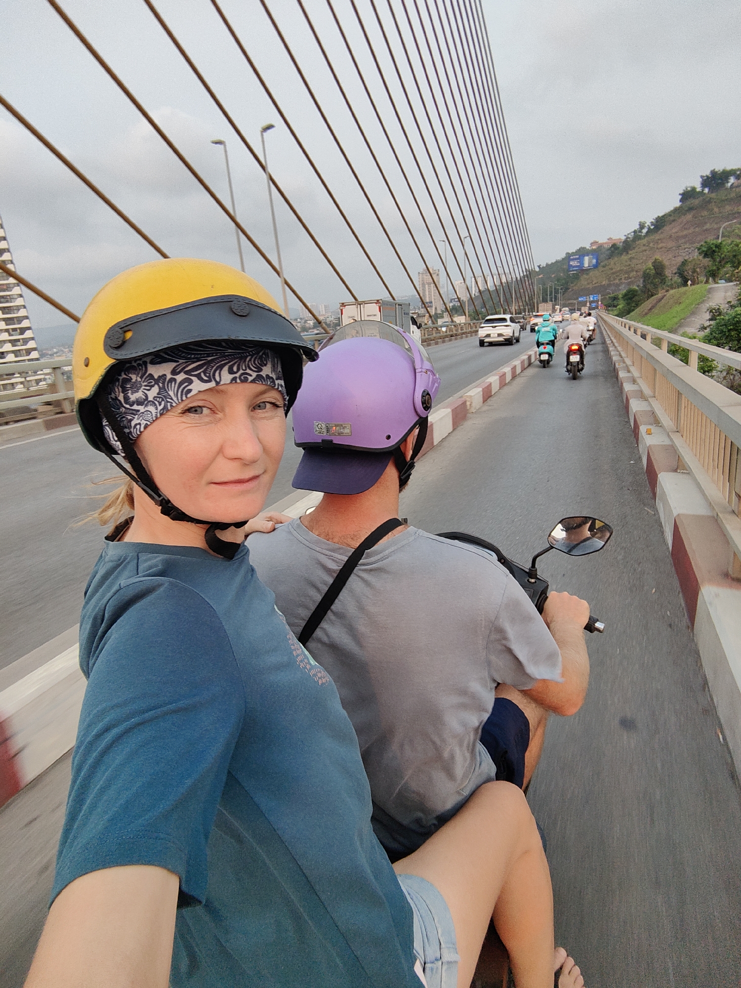 A medium, eye-level, outdoor shot shows a woman and a man riding a scooter on the Bai Chay Bridge in the city of Hong Gai Ward, Vietnam. The woman, seated behind the driver, is on the left side of the image. She wears a yellow helmet over a dark blue paisley bandana, a teal t-shirt, and denim shorts, and she is looking towards the camera. The man is wearing a lavender helmet, a gray t-shirt, and shorts. His back is to the camera. Both riders are white and fair-skinned. A strap for a bag is across his back. In the background is traffic on the bridge, which includes cars and scooters. A light gray sky with many cables from the bridge. A hillside and a building are visible in the background. The railing of the bridge is a light peach color with a wide red and white stripe underneath. The scooter’s side mirror is in the frame on the right. The woman’s bare foot is visible near the bottom right.