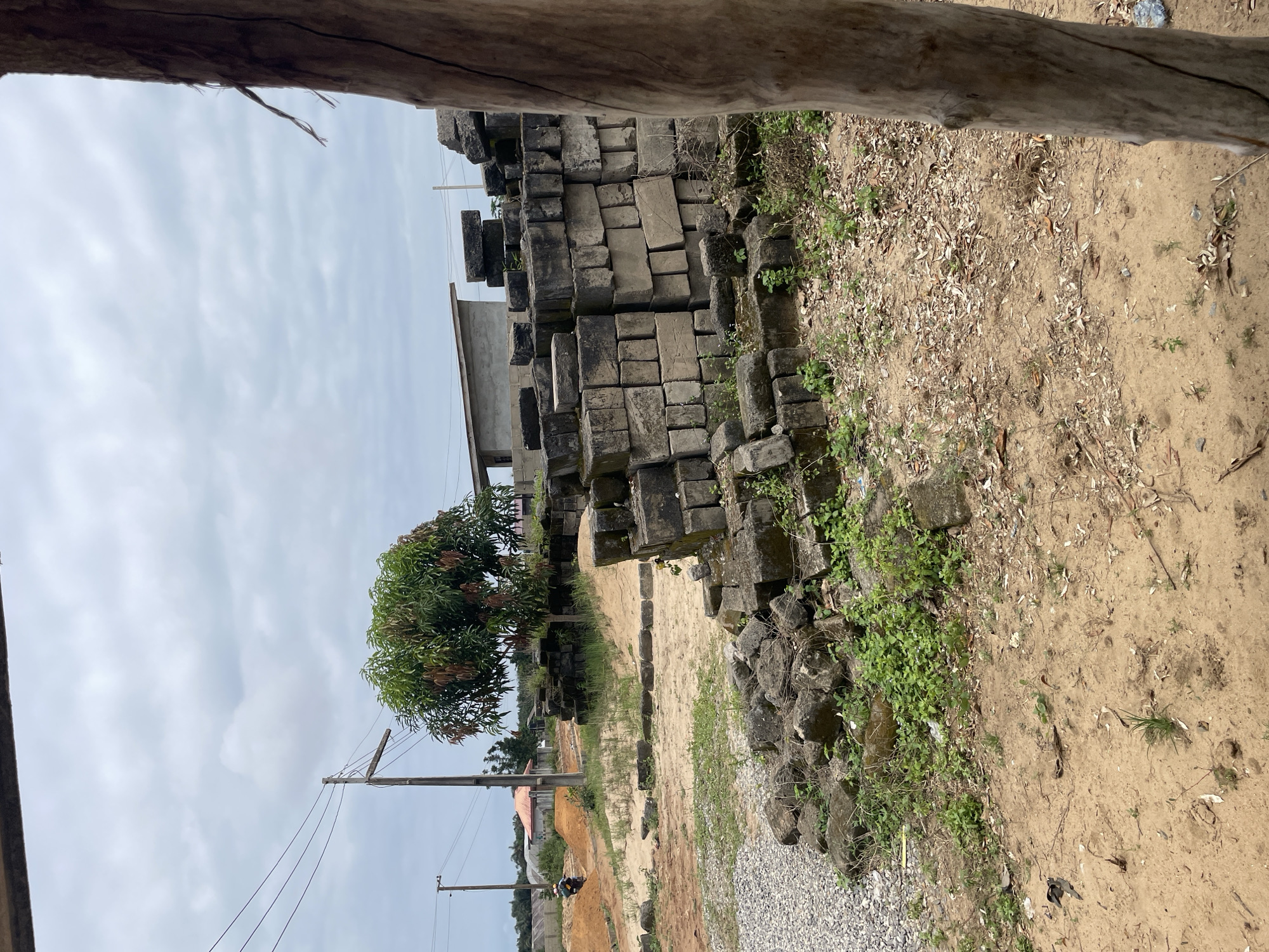 The image depicts an outdoor scene in Iyafin, Nigeria, with a focus on construction materials and the surrounding environment. In the foreground, a pile of weathered concrete blocks is scattered on the sandy ground, interspersed with sparse green vegetation. To the left of the blocks, a gravel pile is visible. Further into the image, a dirt road curves, and a person on a motorcycle is traveling away from the viewer. Beyond the road, a cluster of buildings with varying rooflines and construction stages are present. A prominent mango tree stands between the buildings and the piles of concrete blocks. Utility poles with electrical wires traverse the sky, and the sky itself is overcast with scattered clouds. A large, textured wooden post is prominently featured on the right side of the frame, partially obscuring the view. The overall impression is a construction or undeveloped area within a town or village setting.