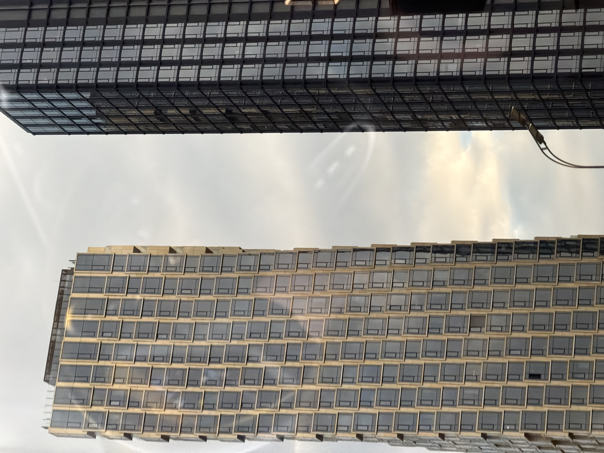 The image shows a view between two buildings, possibly in Moscow, Russia, as indicated by the architectural style often associated with modern buildings in Moscow.
The bottom building appears to have a large facade with many square windows, arranged in a grid-like pattern. The building's exterior has a light, possibly tan, tone. There are reflections in the windows, indicating they are reflective.
Above, another building is visible. It also has many square windows, arranged in a grid pattern. However, the exterior is darker than the building below, possibly black or a very dark gray.
The sky in the background appears to be overcast or hazy, with hints of lighter areas suggesting partial sunshine. There are power lines present in the shot.