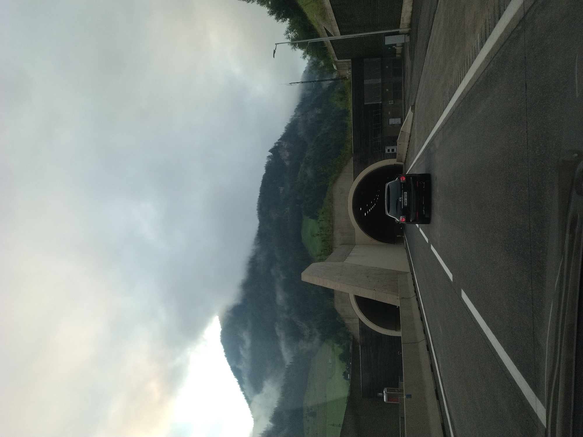 The image captures a view from a vehicle driving on a highway approaching a mountain tunnel in Rothenwand, Austria. The sky is overcast with clouds, and the surrounding mountains are covered in dense green forests, with some areas shrouded in mist. The highway has two lanes in the direction of travel, with white lane markings. In front of the vehicle, a dark-colored SUV is driving towards the tunnel entrance. The tunnel entrance is a large, arched opening in the mountainside, with concrete structures surrounding it. To the right of the road, a street lamp is visible. The overall scene suggests a cloudy and possibly damp day in a mountainous region.