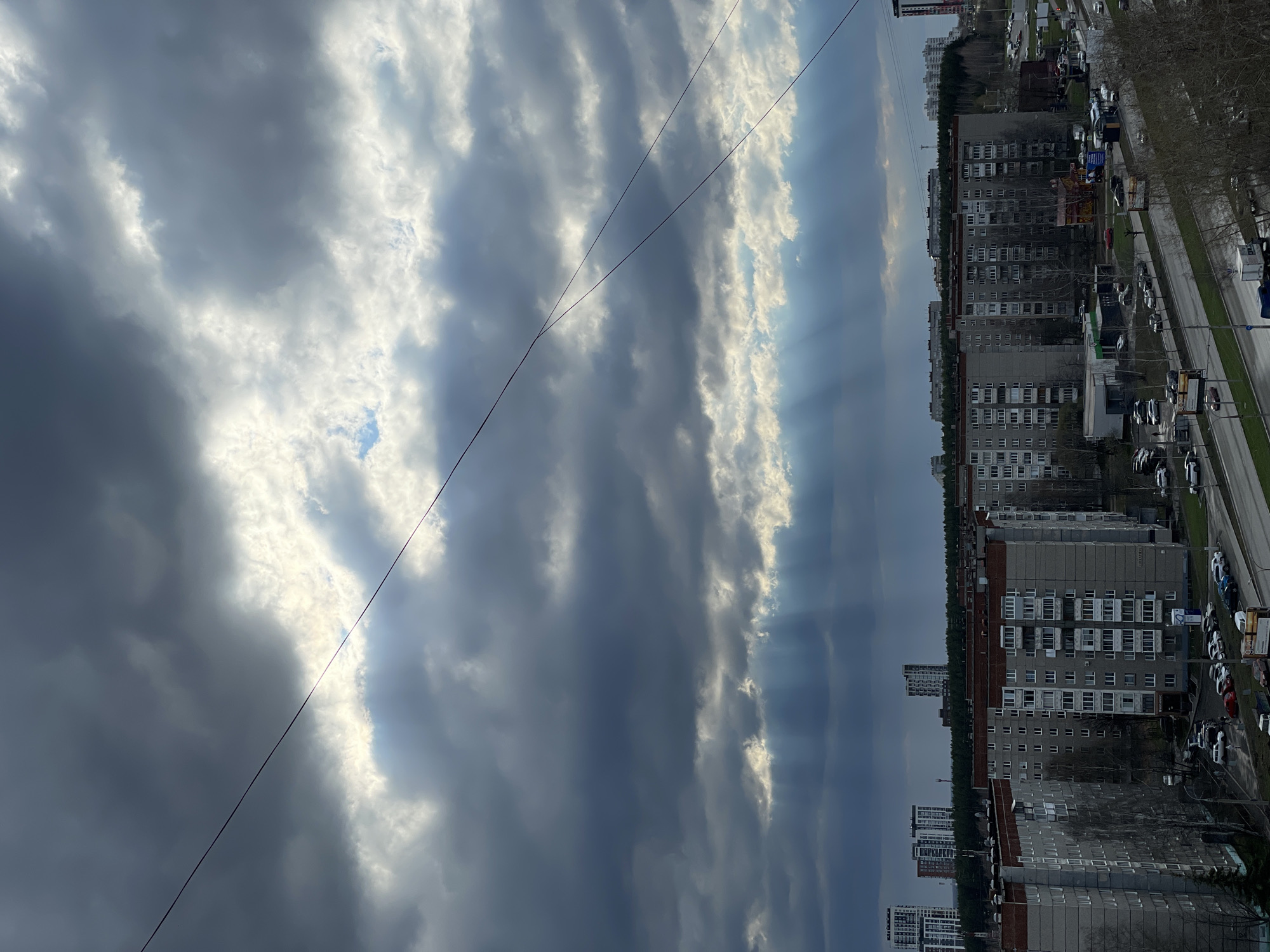 The image captures a wide-angle view of a residential area in Izhevsk, Russia, dominated by a dynamic and cloudy sky. The upper two-thirds of the frame are filled with a complex pattern of clouds, ranging from dark grey to brilliant white, with visible sun rays (crepuscular rays) breaking through gaps on the right side, creating striking shafts of light. A thin, dark power line stretches diagonally across the sky, from the lower-left to the upper-right.
Below the horizon, an urban landscape unfolds. Several large, multi-story apartment buildings, characteristic of Soviet-era architecture with their grey facades and repetitive window structures, line a wide street. A continuous line of trees, mostly bare, runs behind the main row of buildings, and additional high-rise structures are visible in the distance, suggesting a dense urban environment. Numerous cars are parked along the street, and others are visible in motion, indicating active daily life.
The time of day appears to be daytime, possibly late afternoon, given the dramatic lighting from the sun breaking through the clouds. The weather is partly cloudy to overcast. The scene depicts a typical city residential district. While no people are distinctly visible, the presence of vehicles and buildings suggests human activity. There are some billboards and street signs along the road, but any text on them is too small and blurred to be legible. The overall impression is one of a bustling city residential area framed by a powerful and evocative sky.