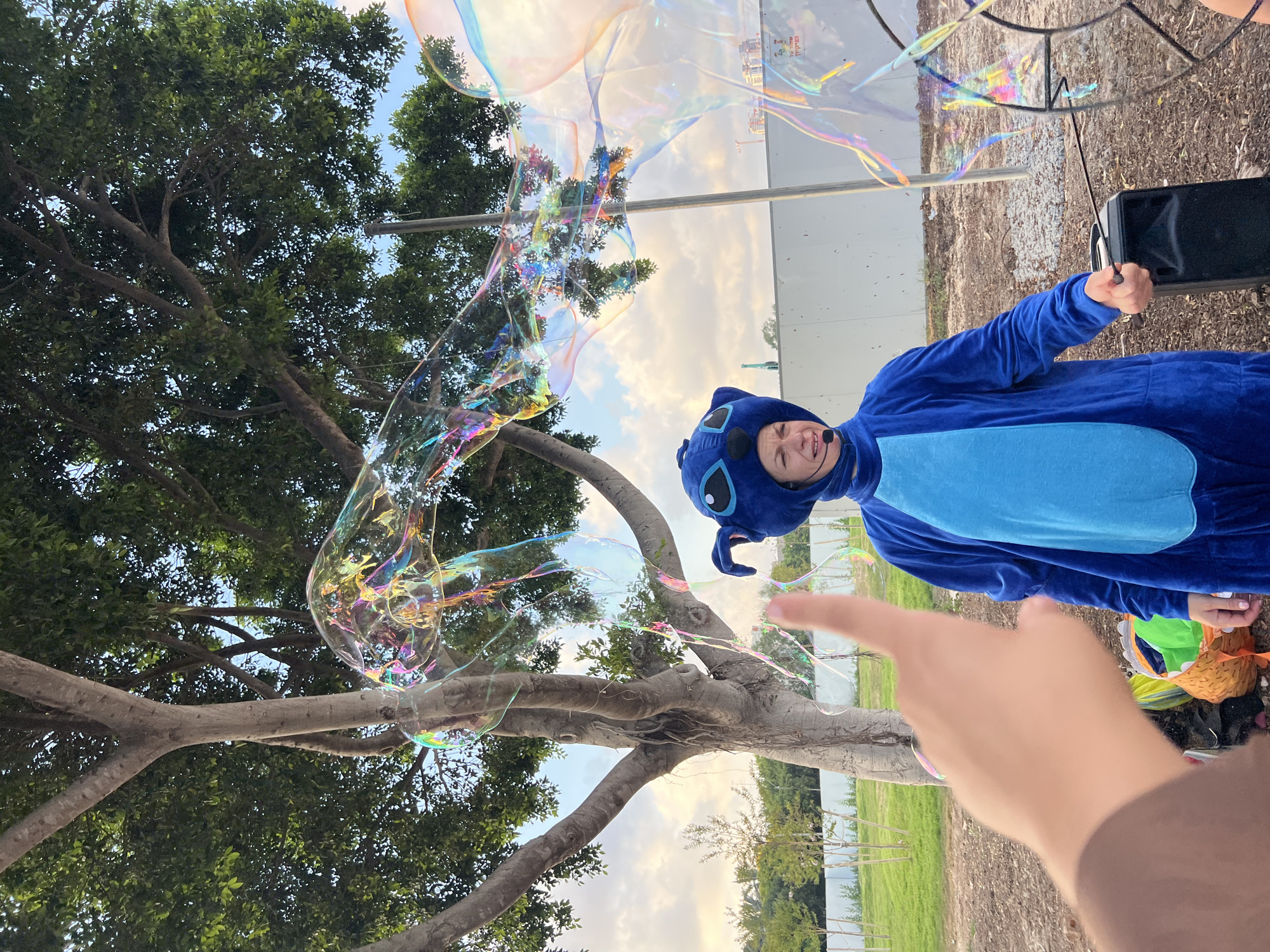 In Kiryat Bialik, Israel, a person in a blue Stitch costume is creating large, iridescent bubbles outdoors. The costume features a hood with Stitch's signature ears and eyes, and a dark blue body with a lighter blue belly. The person wears a headset microphone. A large tree with green foliage is to the left of the frame, and a white fence borders a grassy area in the background. The ground is covered in dirt and gravel. In the foreground, a person's hand is visible, with a finger extended towards the bubbles. Various large, translucent bubbles with rainbow-like reflections drift in the air. One particularly large bubble extends across the upper portion of the image. A black speaker is positioned to the right of the Stitch character. The sky appears overcast with some lighter patches.