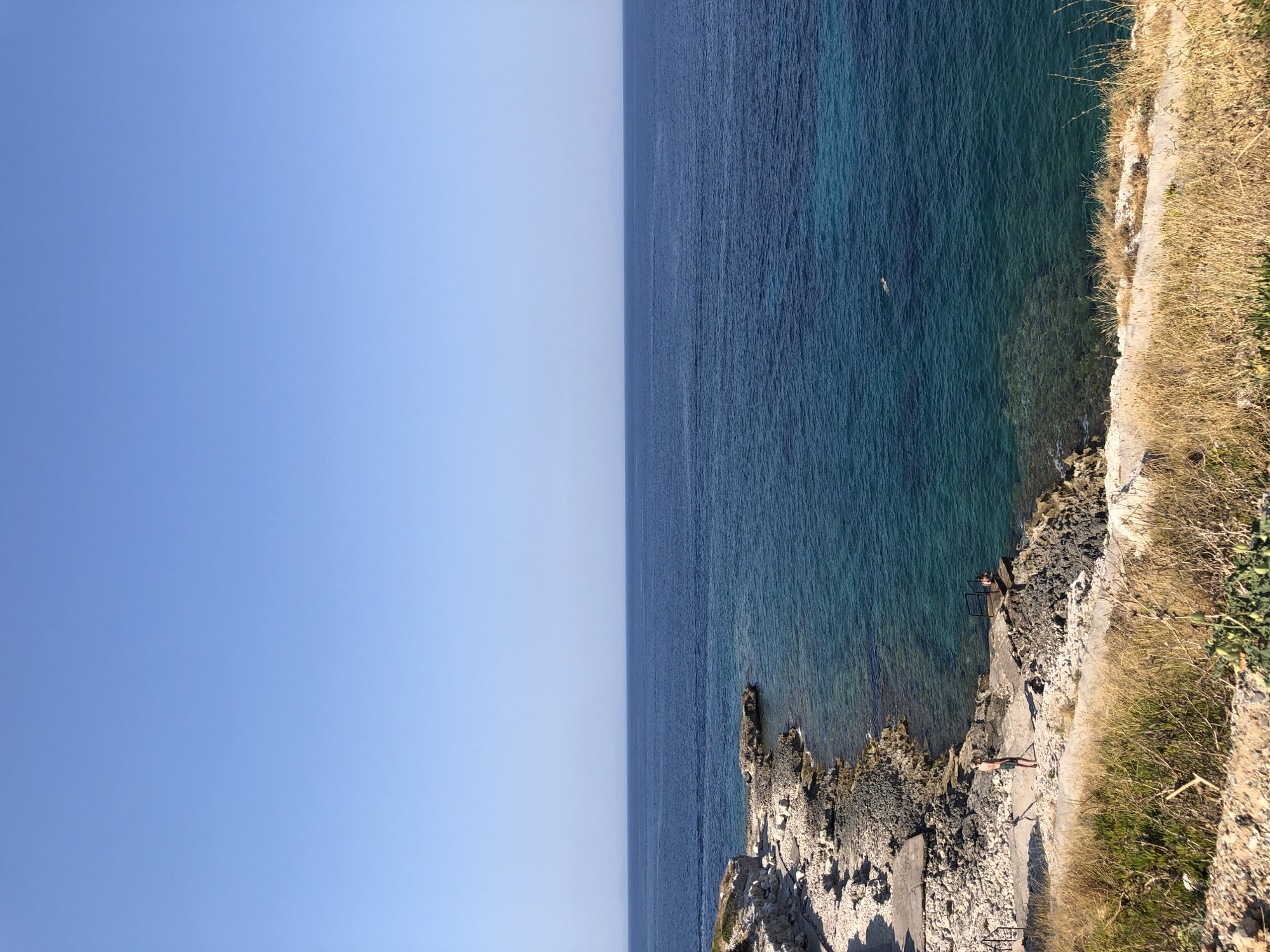 The image depicts a coastal scene on a clear, sunny day. A rugged, rocky shoreline is visible on the left, with a paved pathway leading down to the water. A person stands on the pathway, facing away from the viewer, wearing dark shorts and a lighter-colored shirt or no shirt. Further along the rocky shore, another person appears to be in the water, near a metal railing that leads into the sea. The water itself is a deep blue, transitioning to turquoise near the shore, indicating clear shallows. A single swimmer is visible further out in the darker blue water. The horizon line is sharp, separating the calm sea from a vast, pale blue sky. Dry, golden grass and vegetation line the foreground, suggesting a Mediterranean climate. The location is identified as Karaburun, Turkey.