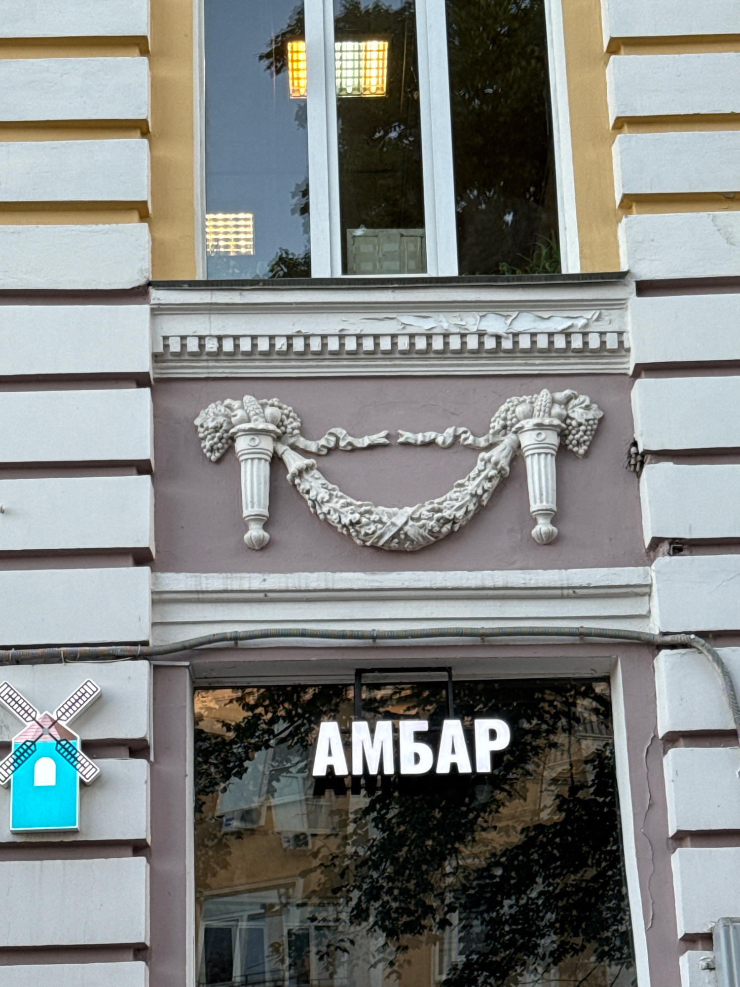 The image shows a close-up of the facade of a building in Tver, Russia. The facade features decorative architectural elements, including a frieze with a garland of fruits and urns. Below this, a large window displays white text on a black background, reading "АМБАР." To the left of the window is a bright blue, stylized windmill-shaped sign. The window reflects the surrounding environment, including trees and other buildings. The overall impression is of a commercial establishment with a classic architectural style.