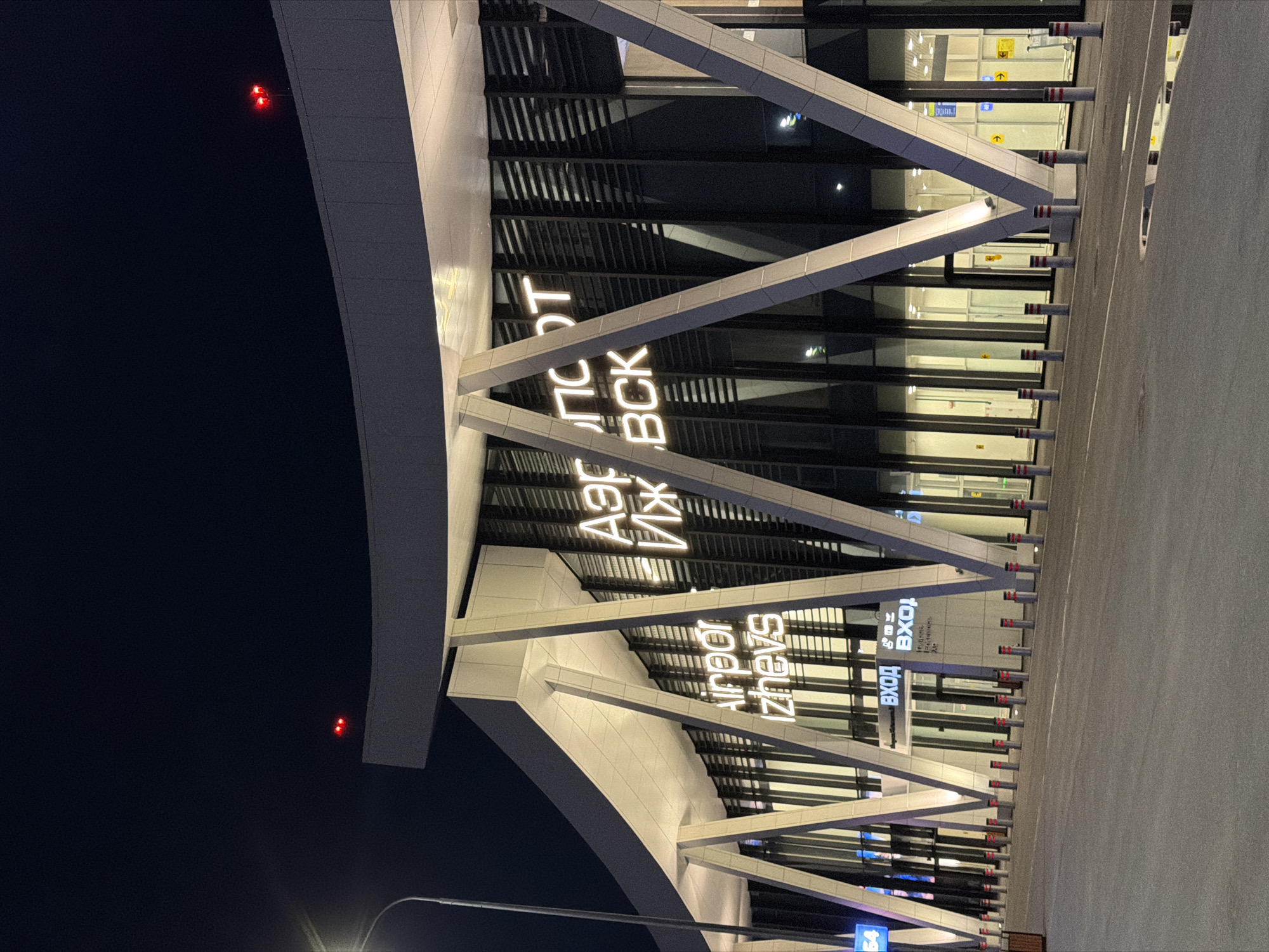 The image depicts the modern facade of Izhevsk Airport, located in Novoye Martyanovo, Russia. The airport building features a contemporary architectural design with a sweeping, curved roof supported by angled white pillars. Large glass windows and dark vertical louvers make up the main body of the building. White neon signs display the airport's name in both Russian and English: "Аэропорт Ижевск" and "Airport Izhevsk."
In front of the building, a paved area stretches out, lined with bollards, some of which have red reflective elements. The lighting suggests it is nighttime or dusk, with the airport's illuminated signage being the primary light source on the building itself. Red lights are visible on the upper edges of the roof, likely navigation or warning lights. The overall impression is of a well-lit, modern transportation hub at night. There are no people visible in the frame.