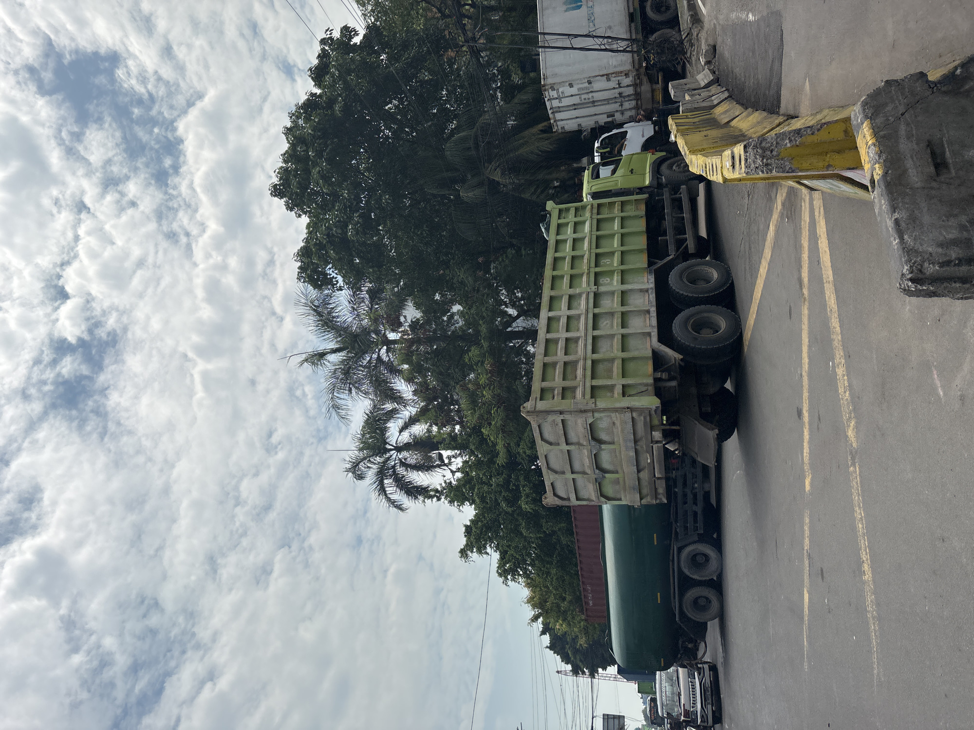 When correctly oriented, the image captures a daytime street scene, likely in an industrial or commercial area of North Jakarta, Indonesia, given the types of vehicles and vegetation. The sky is largely covered with white, fluffy cumulus clouds against a bright blue background, indicating fair weather and clear visibility.
The main subjects are several large trucks occupying the roadway. In the foreground and middle ground, a prominent green dump truck is visible from its side, showcasing its ribbed cargo bed and large wheels. Behind it, a dark green tanker truck is partially seen. Further back, a white container truck is present, with the partial word "LOGIC" or "LOGISTICS" faintly discernible on its side. Other vehicles can be glimpsed in the distance along the road. The number "8" is visible on the side of the green dump truck.
The road features clear yellow markings, including double lines and triangular patterns, suggesting traffic flow or warning zones. To the right, a series of weathered concrete barriers, some painted yellow and others in their original grey, define the edge of the road.
Lush green trees with dense foliage are prominent, with several tall palm trees adding to the tropical setting. Power lines are visible against the sky in the upper parts of the image. The scene depicts a typical busy street, with no specific human activity evident, suggesting either parked vehicles or slow-moving traffic. The time of day appears to be late morning or early afternoon, based on the bright, diffused light.