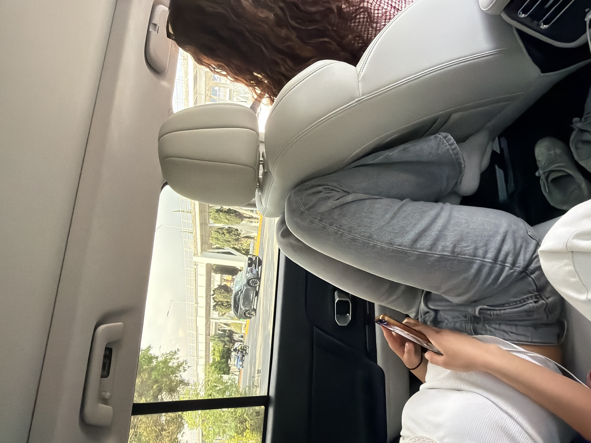The scene depicts the interior of a car, likely in motion, as evidenced by the blurred outside view through the window. The car appears to be traveling along a city street, potentially in Puebla, Mexico, as suggested by the architecture and vegetation visible outside the window. The car's interior is primarily light-colored, featuring white leather seats. A person with long, wavy brown hair is seated in the front, and another person, wearing gray jeans and a white top, is in the back, seemingly occupied with a mobile phone. A pair of sneakers is seen on the car floor.