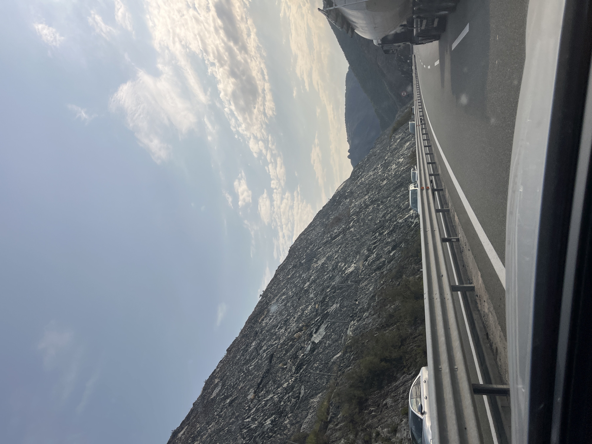The image shows a highway in Rrëshen, Albania, with a steep, rocky mountainside on the left and a valley with more distant mountains on the right. The sky is partly cloudy with a pale blue hue. A metal guardrail separates the highway lanes from the mountainside. Several vehicles are visible on the highway, including a silver sedan on the left, other cars further ahead, and a large tanker truck on the right lane. The road is paved and marked with white lines. The overall impression is a scenic, mountainous landscape with active traffic flow.