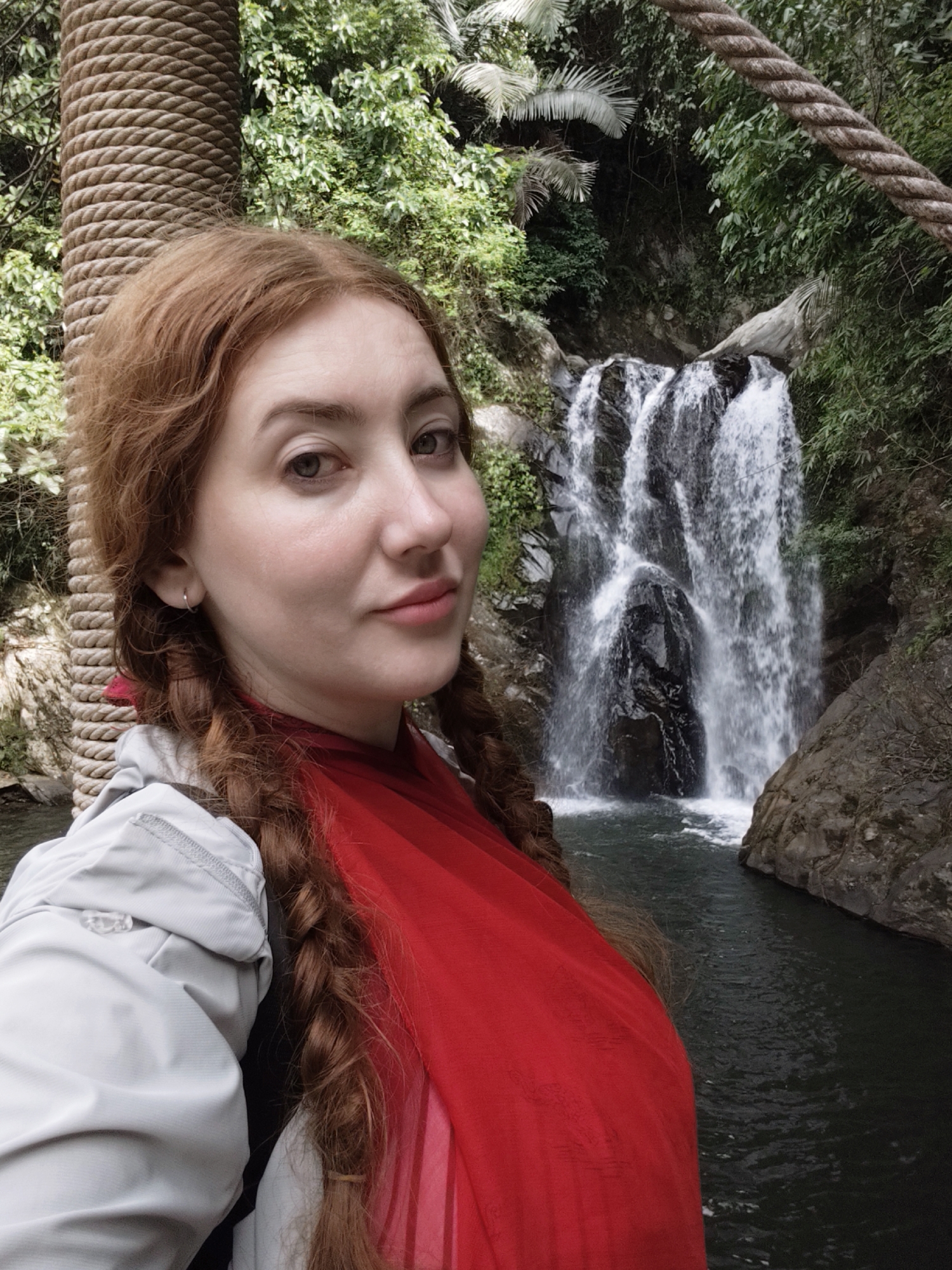 A woman with long, reddish-brown braided hair stands in the foreground of the image, facing the camera. She is wearing a light gray jacket over a bright red scarf or wrap. Her expression is neutral. Behind her, a waterfall cascades down a rocky cliff face into a dark pool of water. Lush green vegetation surrounds the waterfall. Thick ropes and a large, rough-hewn post are visible on the left side of the frame, suggesting the woman may be standing on a bridge or walkway. The lighting suggests it is daytime, likely overcast or in a shaded area.