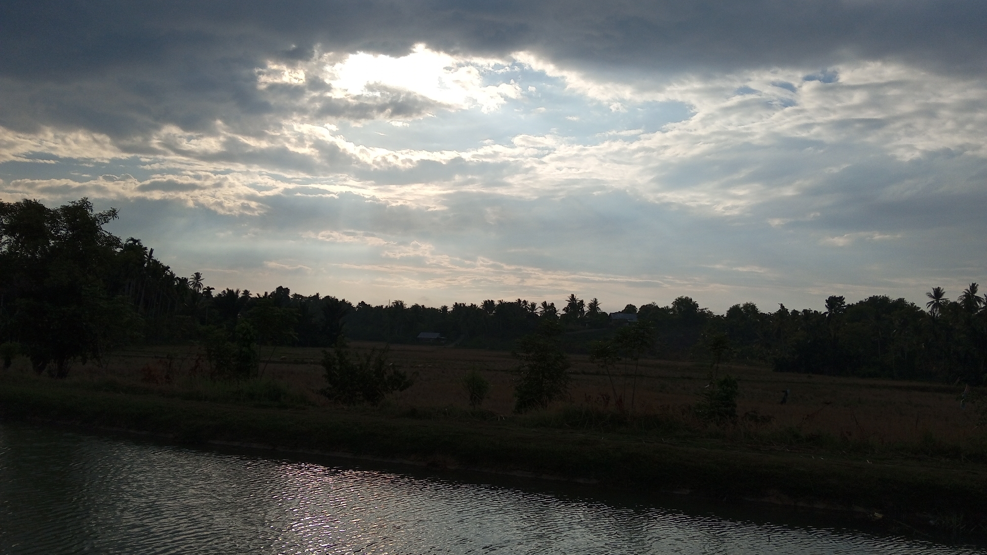 The image depicts a tranquil scene at the edge of a body of water in Buketteukuh, Indonesia. The foreground is dominated by a dark, rippling canal reflecting the overcast sky. The middle ground features a lush, dark green treeline, with hints of harvested fields and a few small structures visible amongst the foliage. In the distance, the sky is a dramatic mix of dark, heavy clouds interspersed with bright patches where sunlight attempts to break through, creating a sense of impending change in the weather. The overall impression is one of natural beauty and the peaceful atmosphere of a rural Indonesian landscape.