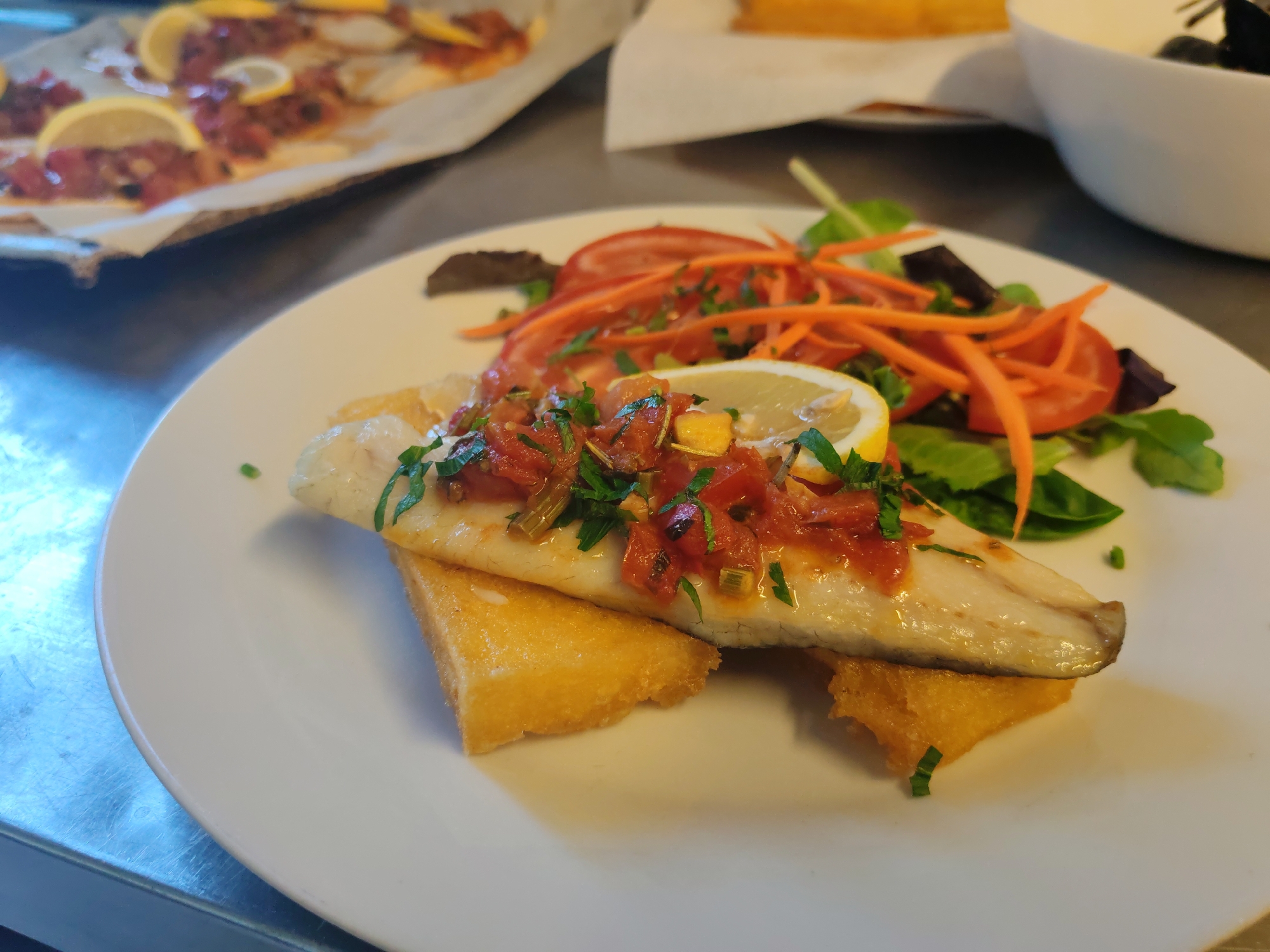A close-up shot shows a white plate with a piece of white fish on top of two pieces of fried polenta. The fish is topped with a red sauce and chopped parsley, and a slice of lemon is placed next to it. A side salad of mixed greens, sliced tomatoes, and shredded carrots is also on the plate. In the background, out of focus, is another plate of food with lemon slices, and a white bowl containing what appears to be black olives. The setting is likely a restaurant in La Spezia, Italy, given the type of dish.
