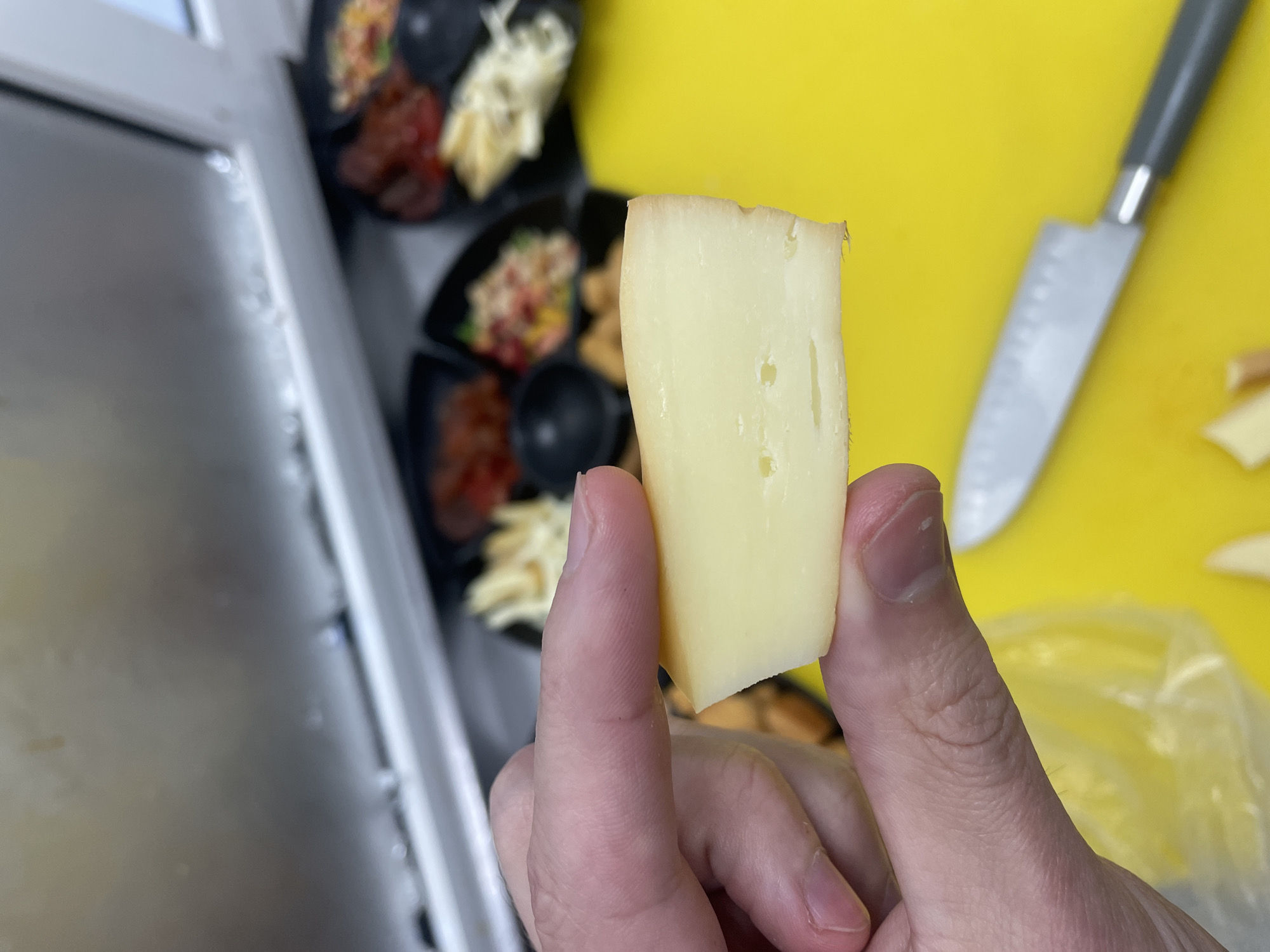 This close-up shot captures a person's right hand holding a rectangular piece of light yellow cheese. The cheese displays a few small, irregular holes or pockmarks on its surface. The hand is seen from the side, with the thumb holding one side and the index finger supporting the other.
The background is softly blurred. To the right, a bright yellow cutting board is visible, featuring a silver knife with a dark handle and some small, lighter-colored pieces, possibly cheese trimmings, near a clear plastic bag. Further back and to the left, several black, compartmentalized dishes hold various out-of-focus food items. These dishes contain what appears to be red chopped ingredients, white shredded food, and other mixed components, suggesting an array of appetizers or ingredients being prepared or served. On the far left, a metallic surface, likely a kitchen counter or equipment, and a white frame are partially visible.
The scene depicts an activity related to food preparation or tasting, likely in a kitchen environment. The lighting is bright and even, indicating an indoor setting. There is no visible text in the image. The location context for this media file is Krasnoyarsk, Russia.