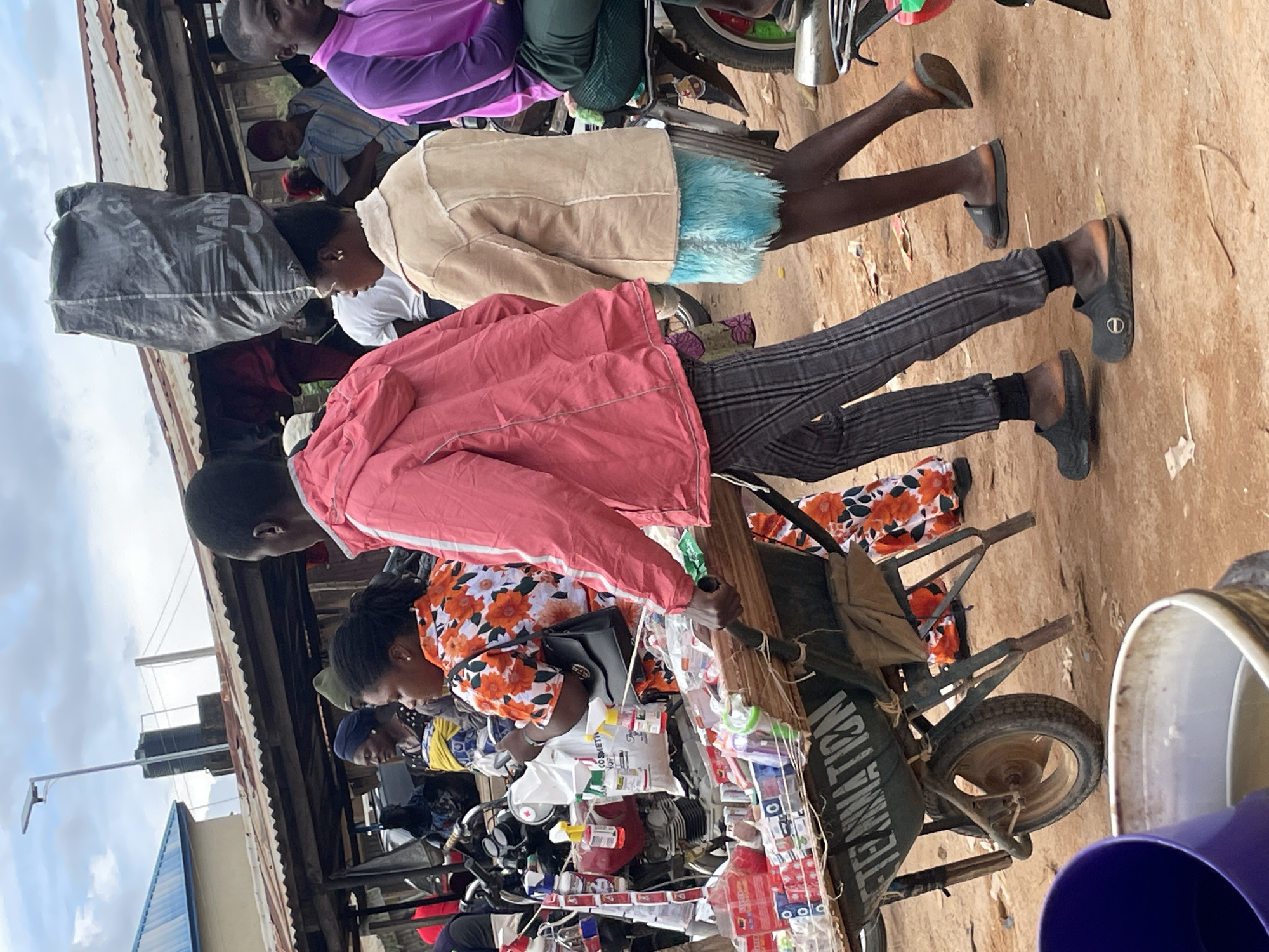 This image shows a street scene in Vom, Nigeria. Several people are visible, mostly women, dressed in casual clothing. Some are wearing bright, patterned shirts, while others are in jackets or hoodies. One person is wearing a purple hoodie, another a light brown jacket, and another a pink jacket. They appear to be standing and interacting, possibly at a market stall or in a public gathering.
In the foreground, there is a wheelbarrow filled with various items, including what appear to be packaged goods like soaps or toiletries, and some drinks. The wheelbarrow itself is dark colored with a visible wheel. Next to the wheelbarrow, there are two large containers, one white and one purple. The ground is dirt and sand, typical of an outdoor setting. In the background, there are structures that suggest a market or informal settlement, with corrugated metal roofing visible. Power lines and a street lamp can also be seen in the distance, along with a tall water tower.
The overall impression is of a busy, everyday scene in a Nigerian community. The lighting suggests it is daytime, likely with cloudy skies given the diffuse light. There are no strong indicators of specific emotions, but the scene appears to be one of normal daily activity. Some text is visible on the wheelbarrow, which reads "TEAMNATION".