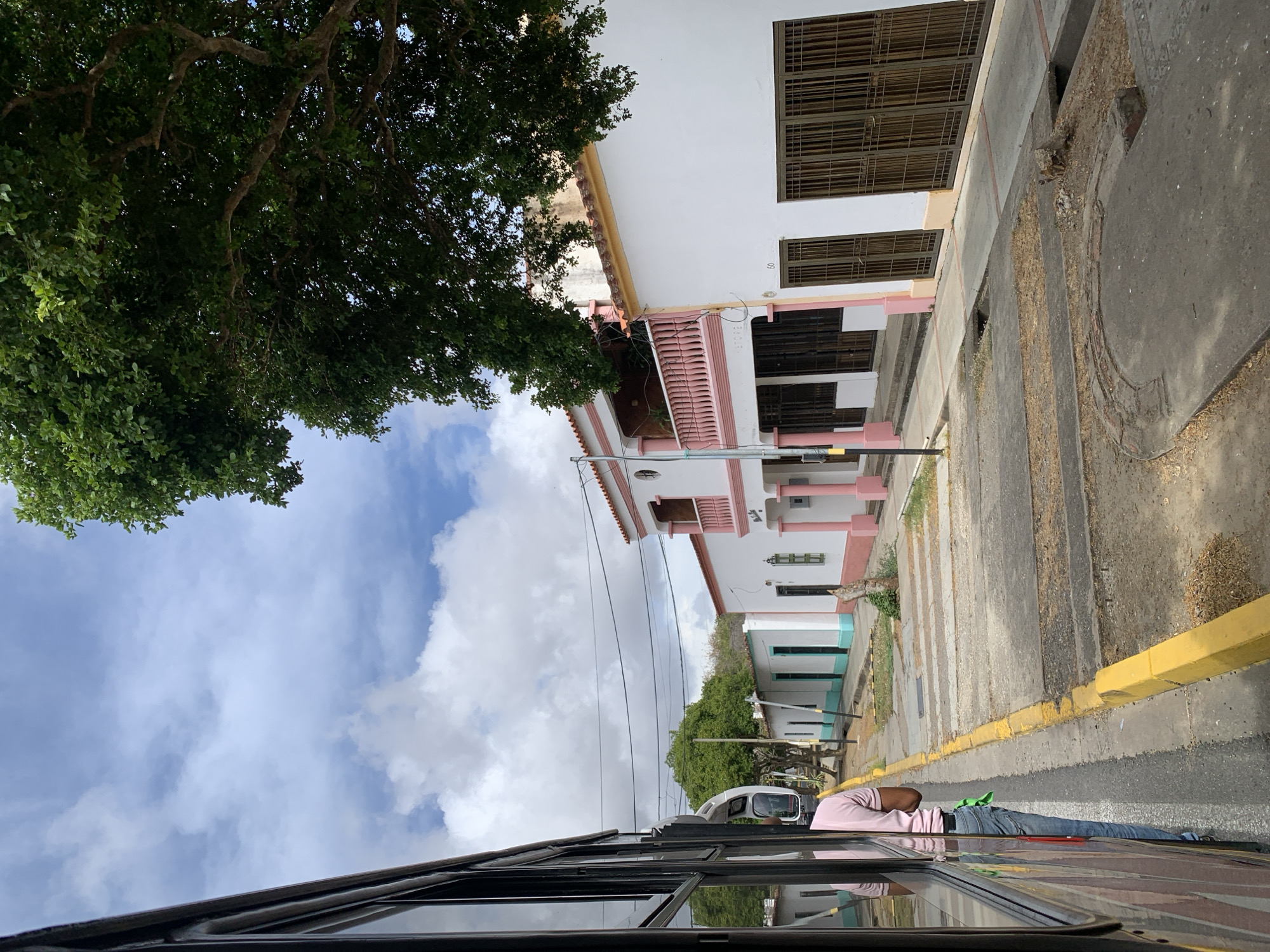 The image, likely taken from inside a moving vehicle, shows a street scene in Santa Ana, Venezuela. A man lies prone on the sidewalk with his head near the curb, wearing a light pink shirt and blue jeans. The curb is painted yellow. The road is visible, with a crosswalk painted on the pavement. The buildings lining the street are multi-story and have architectural details such as balconies and window bars. A tree is visible in the upper left corner of the frame with the sky and clouds above. The vehicle the image was taken from has a dark-framed window.