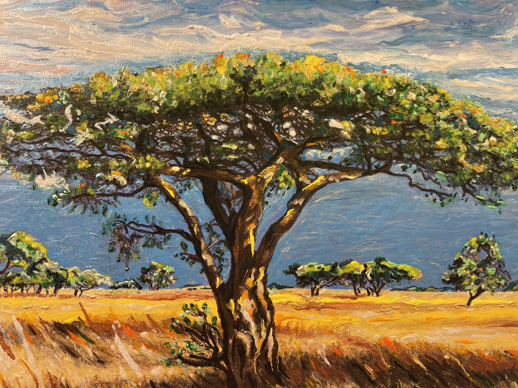 The image is an oil painting depicting an African savanna landscape. A large, solitary acacia tree with a wide canopy dominates the foreground. Its trunk is dark brown with patches of yellow and white highlights, and its branches spread out to form a dense green foliage. Scattered in the background are smaller trees and shrubs, also with green foliage. The ground is covered in dry, yellow grass, with some hints of orange and brown. The sky is a vibrant blue with textured, white and pinkish clouds. The painting style is impressionistic, with visible brushstrokes and thick impasto. The location is not specified within the city of Minsk, Belarus, as the scene is a natural landscape characteristic of Africa. There are no people or animals depicted in the painting.