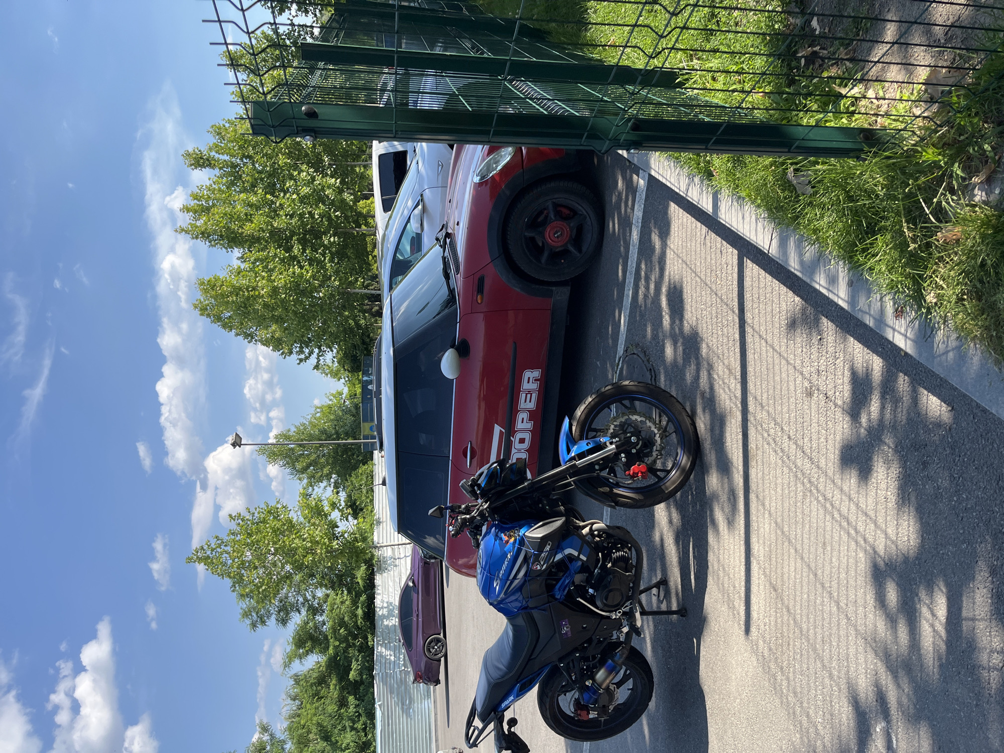 A blue and black sport motorcycle is parked on an asphalt lot next to a red Mini Cooper car with "COOPER" written in white letters on its side. Behind the Mini Cooper, a white SUV is visible. Further in the background, a purple sports car is parked near a metal fence and a row of green trees. The sky is blue with scattered white clouds. The scene appears to be a parking lot on a sunny day in Dnipro, Ukraine. The image is framed by a green metal fence on the right side.