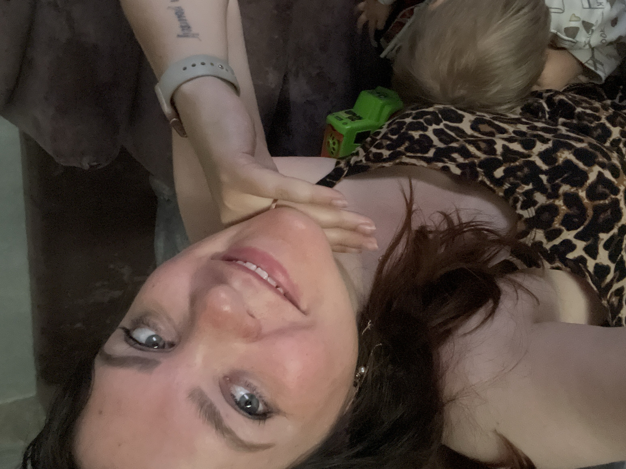 In a close-up, overhead shot, a woman with brown hair and blue eyes is looking at the camera, smiling. She is wearing a leopard print top and a grey smartwatch. Her left arm is resting on her chin, and her right arm is tucked behind her. A small child with blonde hair is sleeping on her chest, their head resting on her shoulder. A green toy truck is visible near the child's head. The background is dark and appears to be a couch. The image appears to have been taken indoors in a residential setting. There is no visible information to suggest the location is in Noviy, Russia.