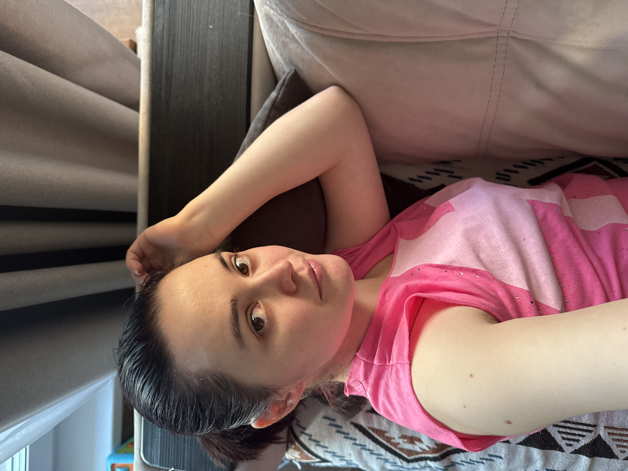 A woman is lying on a couch. She is wearing a pink and white shirt. Her hair is dark and slicked back. The couch has a patterned throw blanket on it. The couch is located in Izhevsk, Russia.