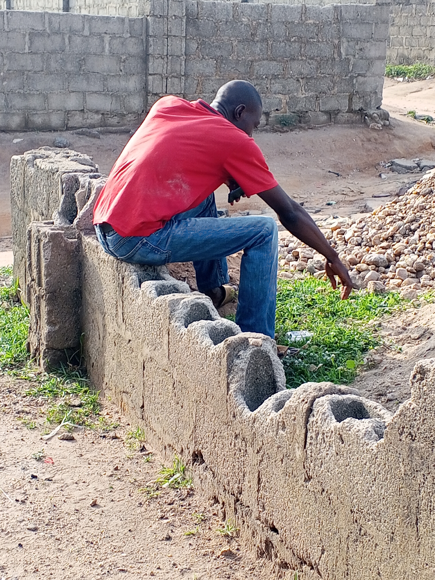 This eye-level, outdoor shot depicts a man in a semi-developed or construction area, likely in Zango-ndaji, Nigeria, during bright daylight.
The primary subject is an adult male, viewed from behind and slightly to his right. He has dark skin and a bald or closely shaved head. He is wearing a bright red short-sleeved t-shirt, blue jeans, and light-colored sandals. The man is seated on a low, rough concrete block wall in the foreground. This wall is constructed from hollow concrete blocks, exhibiting an unfinished or utilitarian texture.
He is leaning forward, his attention directed towards the ground in front of him. His right arm is extended downwards, fingers splayed, as if he is touching, examining, or interacting with a small patch of green grass and weeds growing on the sandy terrain. A small, light-colored cylindrical object, possibly a discarded can, is visible within this patch of greenery. In his left hand, positioned near his lap, he holds a dark, elongated object, which could be a mobile phone or a small tool.
The background features a taller, grey concrete block wall, suggesting ongoing construction or property demarcation. To the man's right, a considerable pile of small stones or gravel lies on the sandy ground. The overall setting is characterized by dusty, sandy earth with scattered tufts of green vegetation, indicative of a rustic, outdoor environment. There is no visible text in the image.