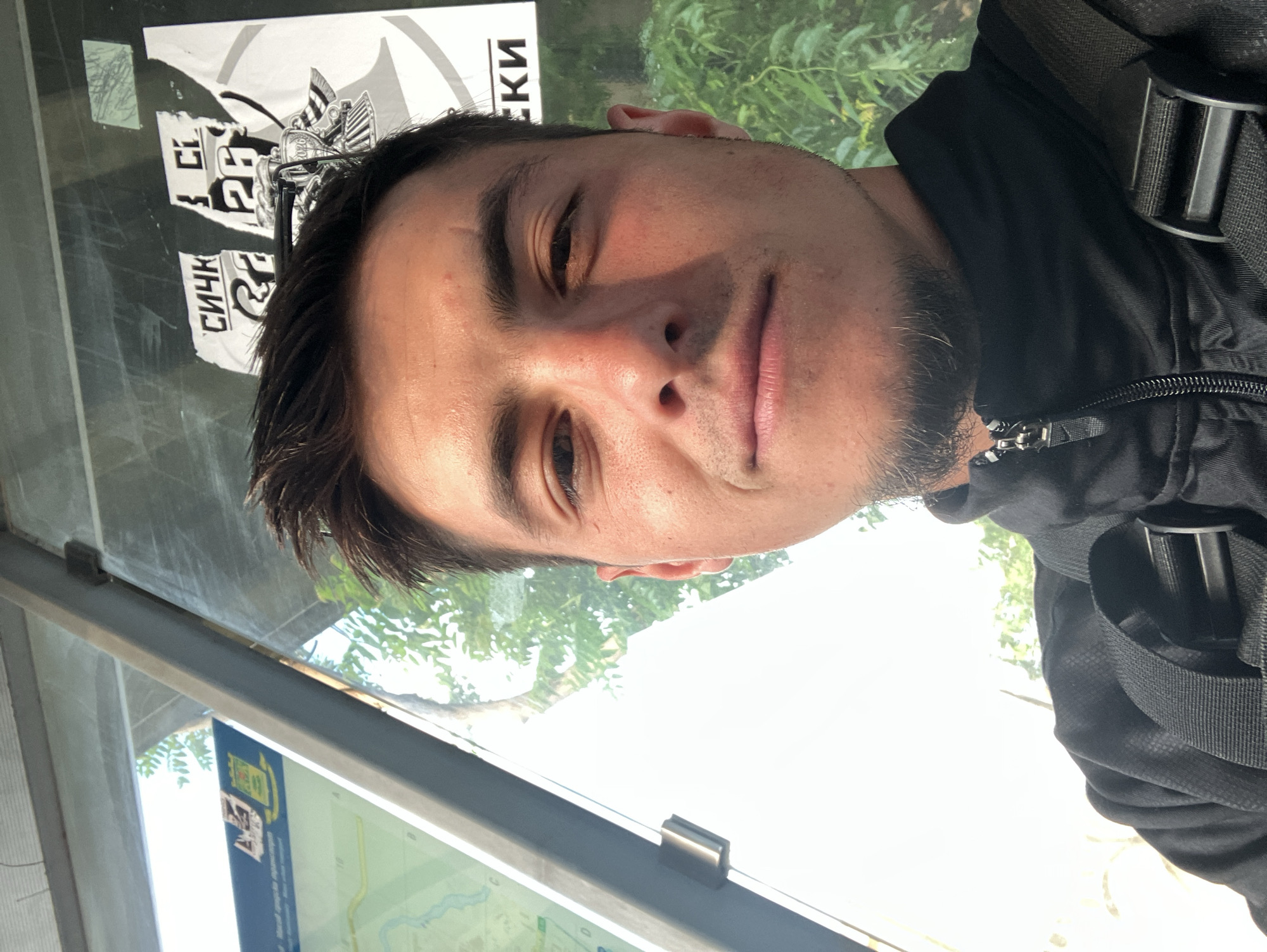 A medium shot depicts a man at what appears to be a bus stop in Plovdiv, Bulgaria. He is fair-skinned, with dark hair, stubble, and visible pores. The man wears a dark-colored jacket with zipper and a black backpack. He is positioned close to a public transport schedule. The background includes posters with partially legible Cyrillic text and green foliage seen through glass. The light suggests an outdoor, daylight setting.