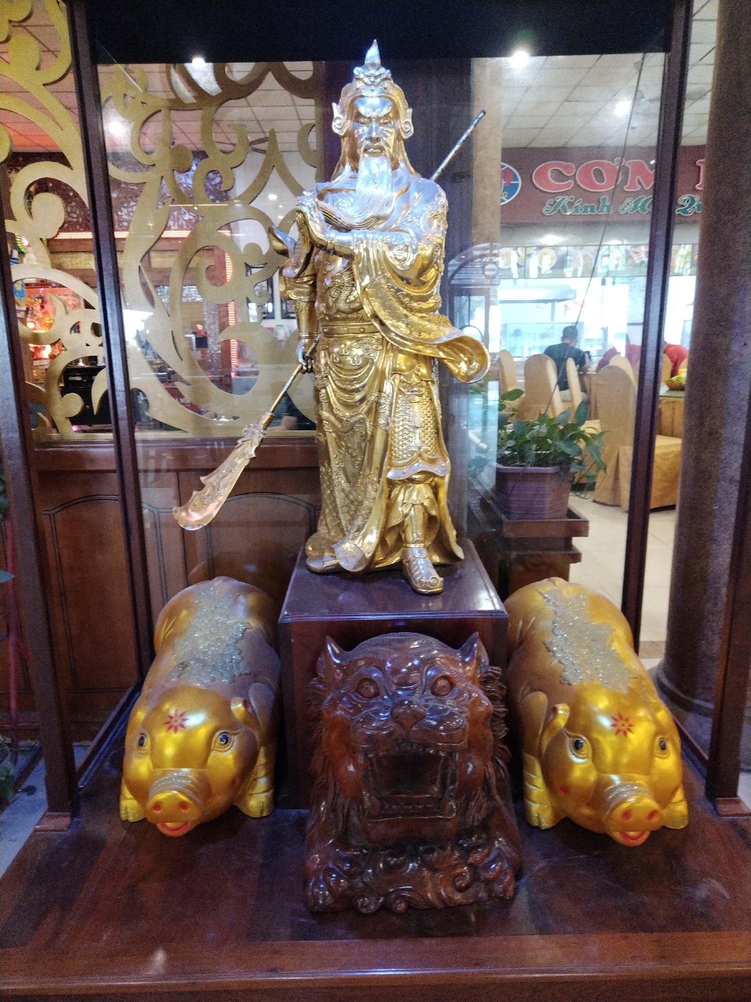 A display case containing a golden statue of a Chinese deity is flanked by two golden pig figurines and a wooden tiger figurine. The statue stands on a raised platform within the case, holding a weapon. The pig figurines are gold with reddish-pink noses. The tiger figurine is carved from wood and has an open mouth, as if roaring. The display case is made of wood with glass sides. Reflections in the glass show a restaurant interior with tables, chairs, and people. The location is in Chu Văn An Ward, Vietnam, as identified from the restaurant sign in the reflection.