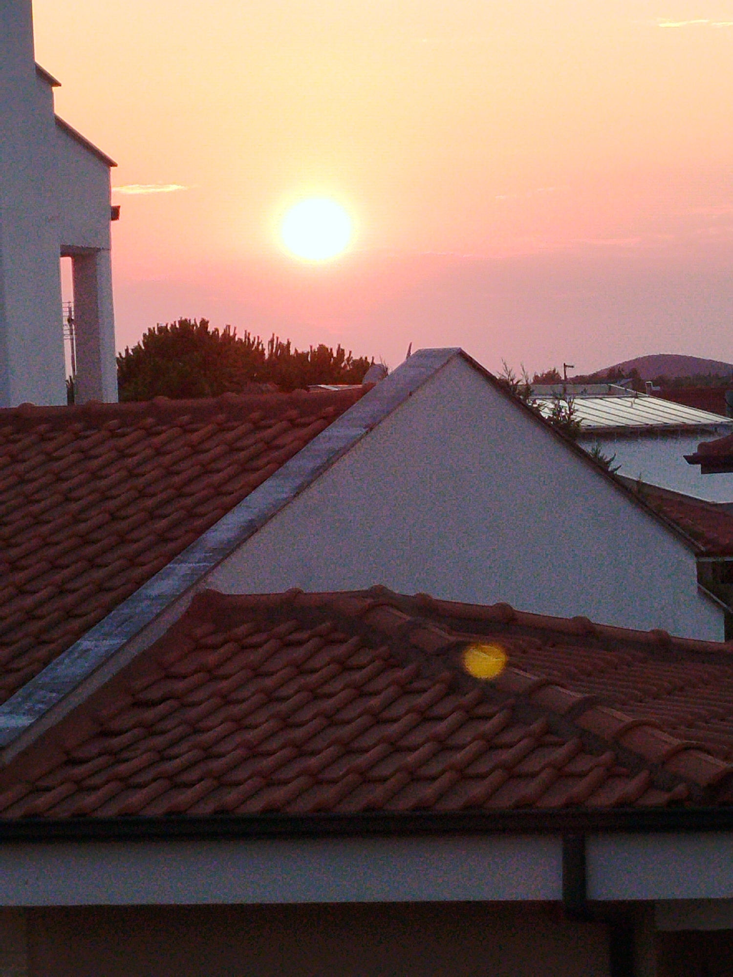 The image depicts a sunset over a residential area in Sithonia Municipal Unit, Greece. The sky is a gradient of orange and pink, with the sun a bright, glowing orb. In the foreground, we see several terracotta-tiled roofs of houses. A white building with a visible opening is on the left side of the image. Behind the roofs, trees and other buildings are visible, with a hillside in the distance. The overall impression is a peaceful evening scene.