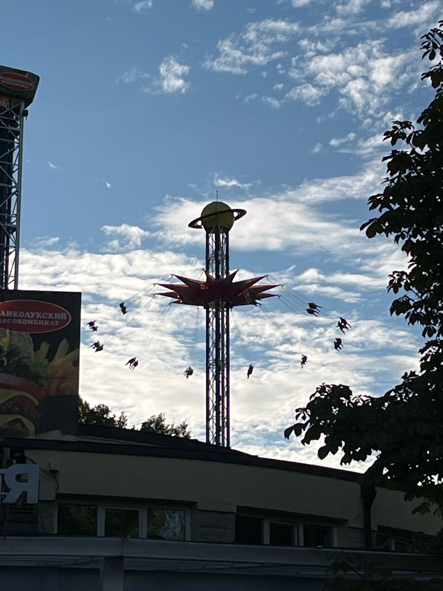 The image shows a tall amusement park ride resembling a swing carousel against a bright blue sky with scattered white clouds. The ride is a towering structure with a central column from which numerous swings hang, each occupied by at least one person. The swings are in motion, creating a circular pattern against the sky. The top of the ride is crowned with a yellow spherical object with a ring, resembling a planet. To the left of the ride, a portion of a tall, metallic structure with a sign is visible. The sign has Russian text and an image of a hamburger and fries. In the foreground, the roofline of a building with windows is visible, partially silhouetted against the sky. A dark, leafy tree branch extends into the frame from the right side, obscuring part of the sky and ride. The scene appears to be an outdoor amusement park, likely during daylight hours. The presence of Russian text on the sign suggests the location is in Russia. Without further information, the specific location within Saint Petersburg cannot be definitively determined from the visual cues alone.