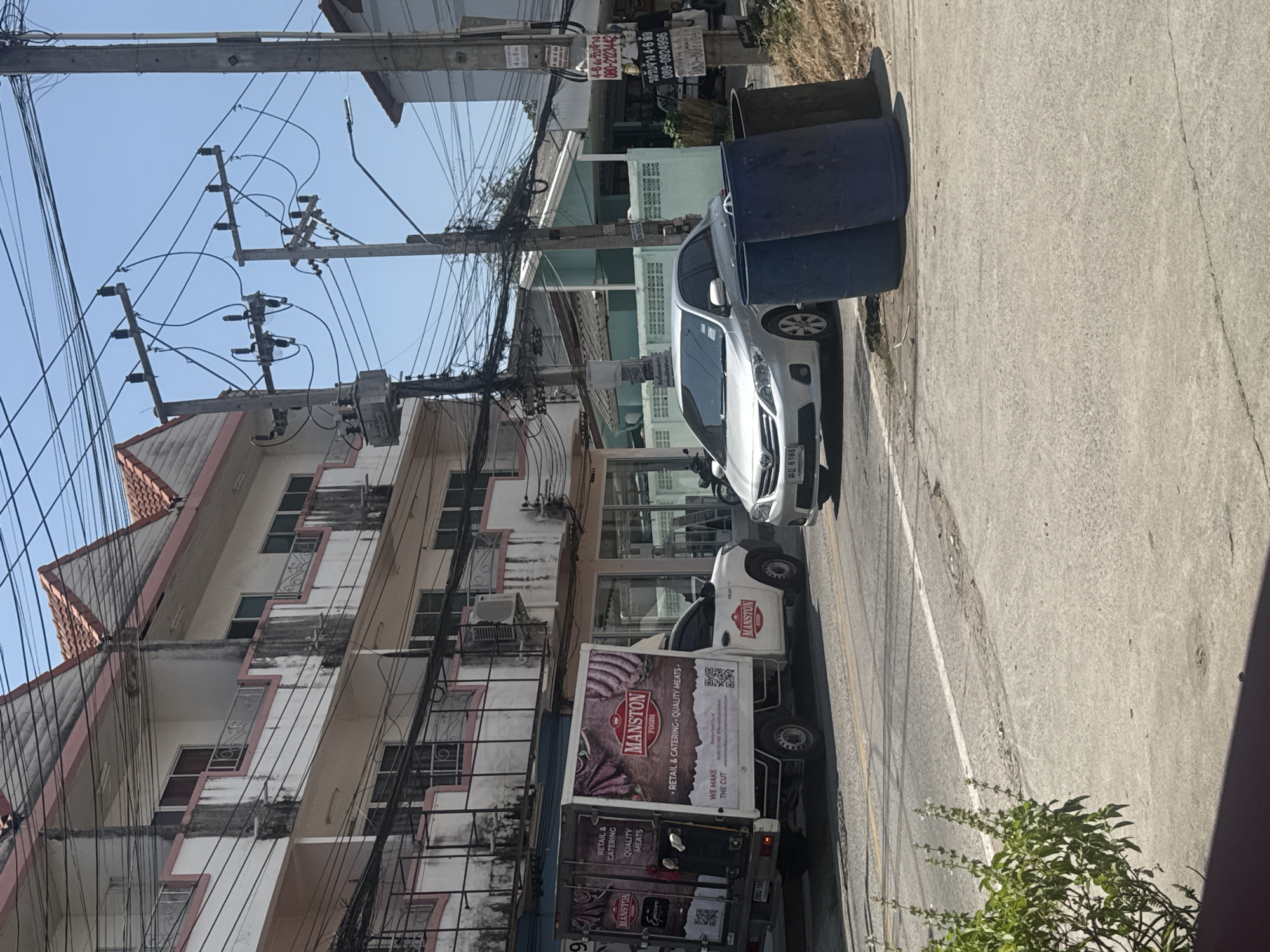This image captures a bright, sunny street scene in Pattaya City, Thailand, originally rotated 90 degrees clockwise. The setting features a bustling urban environment characterized by dense overhead utility lines, local architecture, and parked vehicles.
In the foreground, a concrete road or alleyway stretches across the frame. On the left side of the road, two large, dark blue cylindrical trash bins are placed, followed by a silver car with the license plate "ฎม 6186". Behind the car is a white pickup truck, likely a delivery vehicle, prominently displaying "MANSTON FOODS" branding. The truck also features text like "• RETAIL & CATERING • QUALITY MEATS •" and "WE MAKE THE CUT," along with visible QR codes. A partially obscured Thai license plate (starting with "บส") is visible on the truck.
To the left of the road, multiple multi-story buildings line the street. These structures exhibit local architectural styles, with balconies and visible signs of wear. Numerous utility poles stand tall, heavily laden with intricate and tangled networks of electrical and communication wires, a common sight in the region. Several signs are affixed to these poles, displaying Thai text advertising rental services: "4-6 รับจ้าง 080-2123442" and "รถรับจ้าง 4-6 ล้อ 089-0924996". The sky is clear and bright blue, indicating fair weather. A small patch of green foliage grows on the side of the road in the lower right. No people are visible.