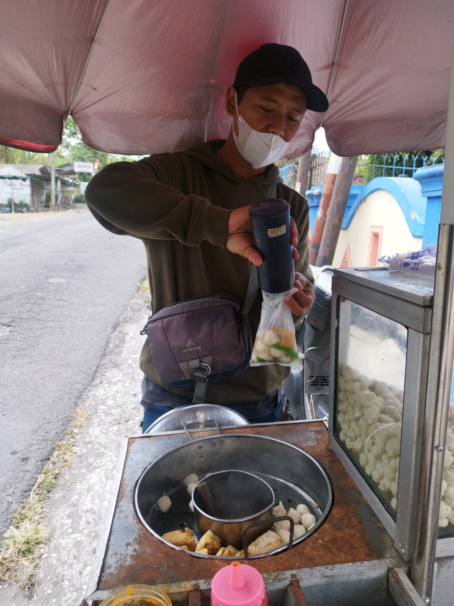 A man wearing a face mask and a black baseball cap is standing at a food cart under a pink umbrella. He is holding a black cylindrical dispenser over a plastic bag filled with meatballs and green chilies, preparing to serve them. The food cart has a metal steaming pot containing meatballs and fried tofu, and a glass display case filled with more meatballs. The scene appears to be on a street in Hargobinangun, Indonesia, with a road and buildings visible in the background.