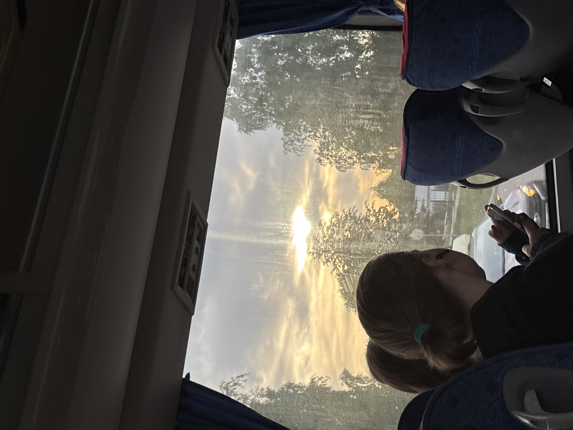 The image shows the interior of a bus, with a young person looking out the window at a sunset. The location appears to be in the city of Chaikovsky, Russia, based on contextual cues. The person is holding a smartphone in their hand and is sitting in a blue bus seat. The view outside the window shows a sunburst with trees and buildings. The image is taken at an angle.