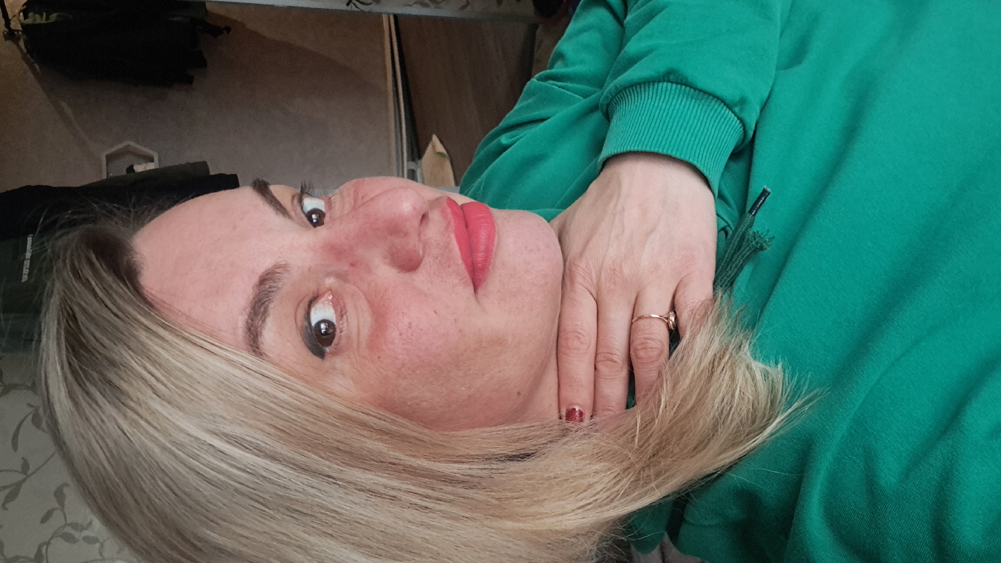 A blonde woman wearing bright red lipstick and a green hoodie is lying on her side. Her hand is positioned near her neck. She is wearing a gold ring on her finger. The location of the photo is unknown.