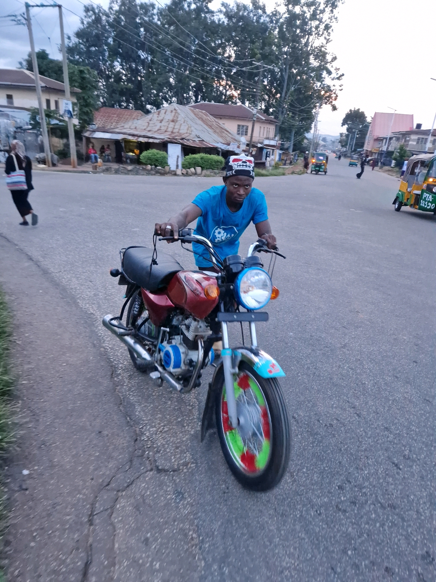 In this medium-shot, eye-level photograph, a young man wearing a blue t-shirt and a black and white skull-patterned cap rides a red motorcycle down a paved road. The man is looking towards the camera with a neutral expression, and his hands are on the handlebars. The motorcycle has a chrome exhaust pipe and a blue accent on the engine. The front wheel is visible, with green and red accents painted on the rim. The background shows a street scene with several buildings, trees, and other vehicles, including a yellow and green auto-rickshaw in the distance. The sky is overcast, suggesting it is either early morning or late afternoon. The environment appears to be a residential or commercial area within the city of Jos, Nigeria, given the presence of the auto-rickshaw, which is a common mode of transport in the region. The road is cracked in several places, indicating it is an older, well-traveled street. A woman in a black headscarf and black pants carrying a striped bag is walking away from the camera on the left side of the street.