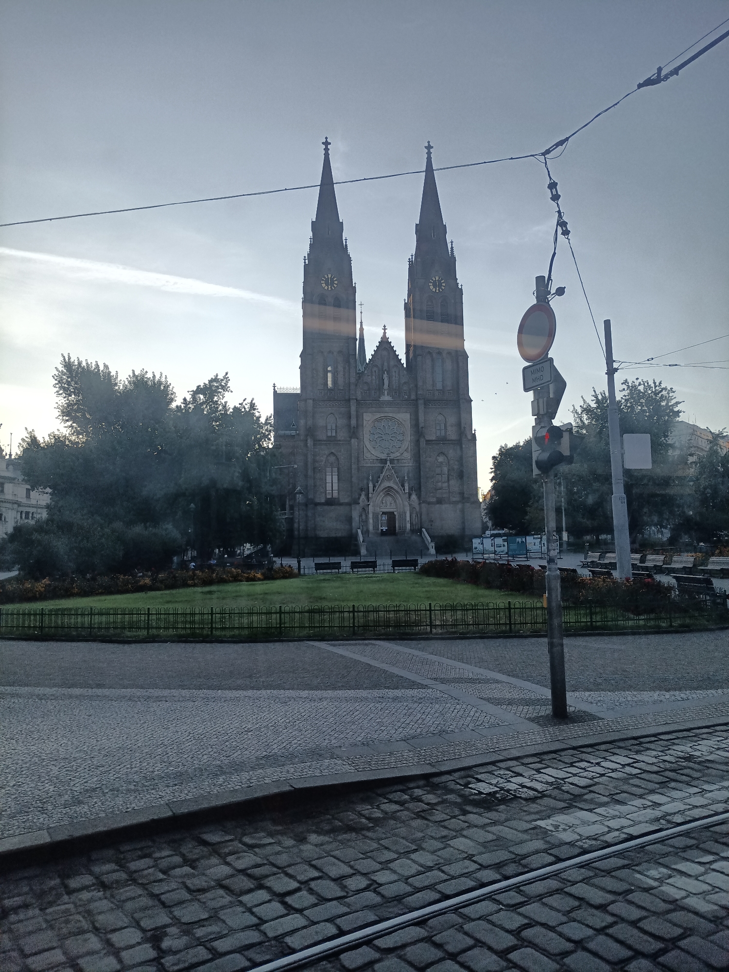 The image captures a view of the Basilica of St. Peter and St. Paul in Vyšehrad, Prague, Czechia, as seen from what appears to be a tram or train. The imposing Gothic Revival basilica dominates the background, with its two tall spires reaching towards a cloudy sky. A large rose window is visible on the facade. In the foreground, cobblestone streets and tram tracks are prominent. A traffic light with a red signal is visible, along with a traffic sign. Trees and a manicured lawn with benches surround the basilica, suggesting a park or public space. The overall scene is muted, with a cool, overcast lighting.